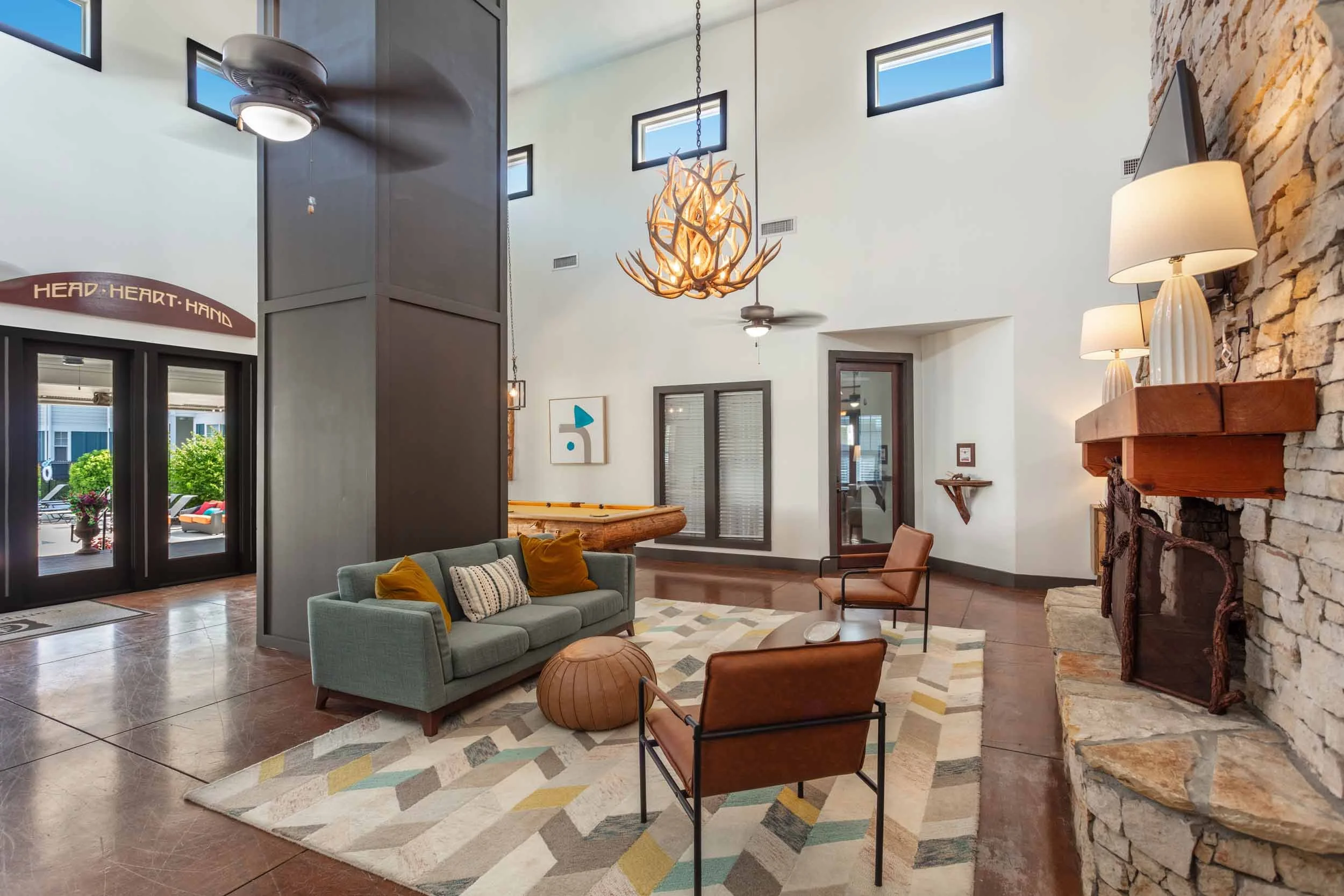  Clubhouse with high ceiling, large wall windows, and a rustic exposed stone fireplace. There is a modern chandelier hanging from the ceiling, and a ceiling fan.