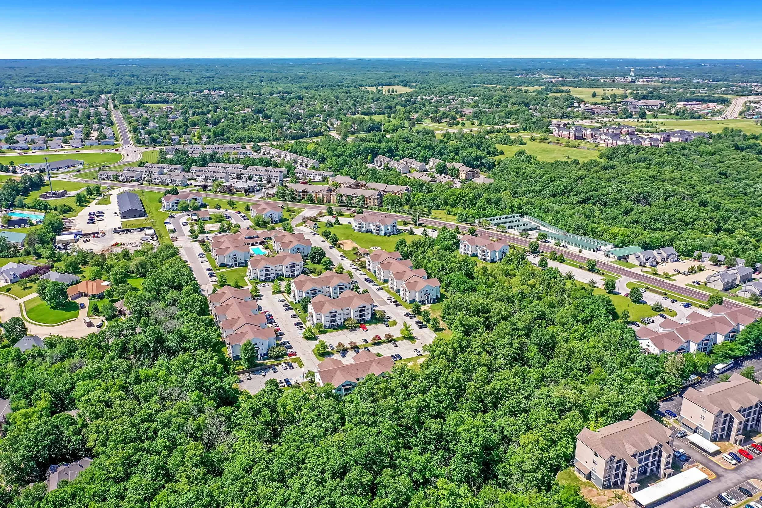 An aerial view of a suburban neighborhood with rows of houses, green trees, and a swimming pool, surrounded by lush forest and open fields under a clear blue sky.