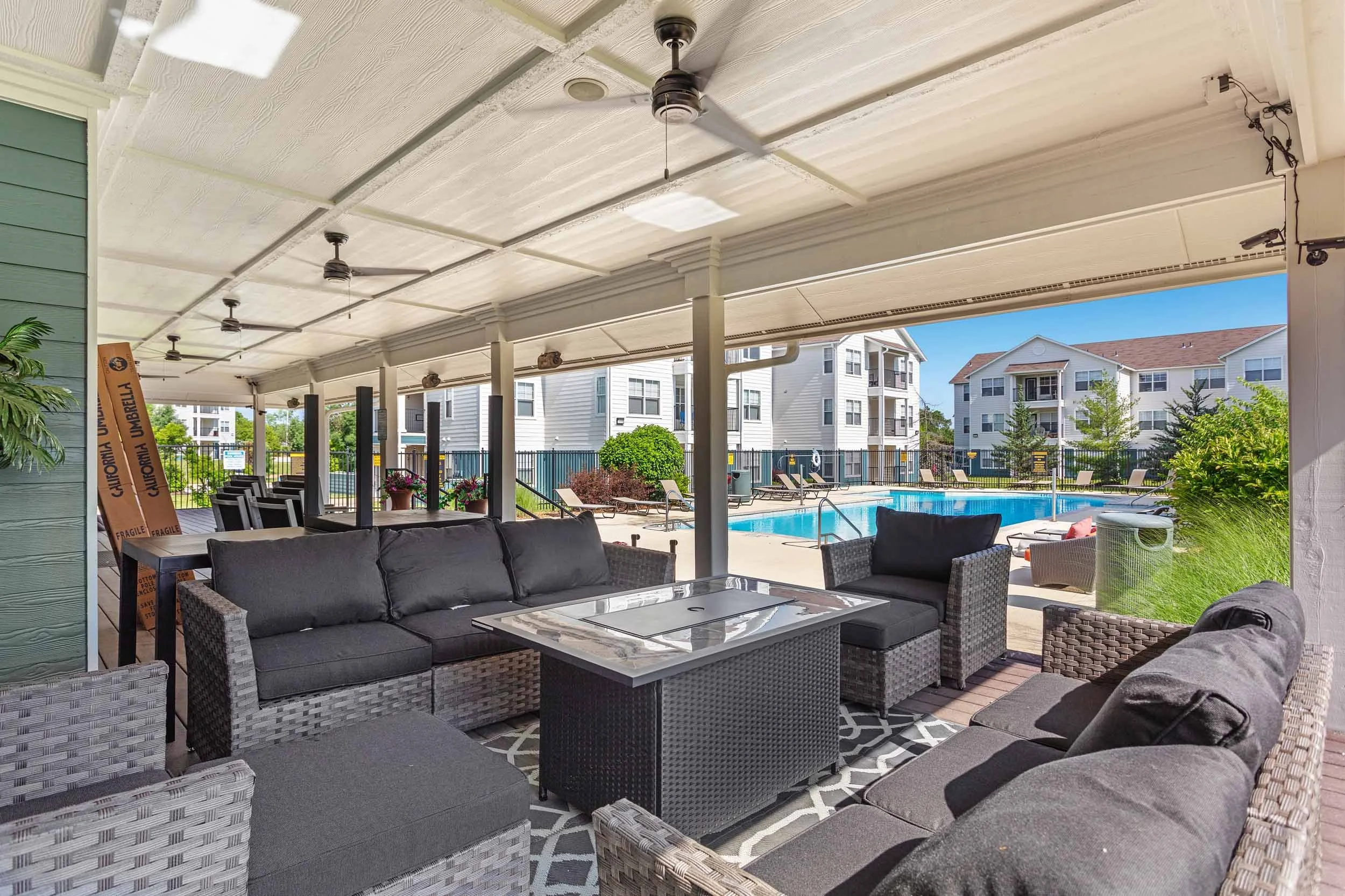 This image shows a covered outdoor seating area with wicker furniture including sofas and chairs, a glass-top coffee table, and a patterned outdoor rug. In the background, there is a swimming pool surrounded by lounge chairs, a fence, and residential buildings, with a clear blue sky overhead.