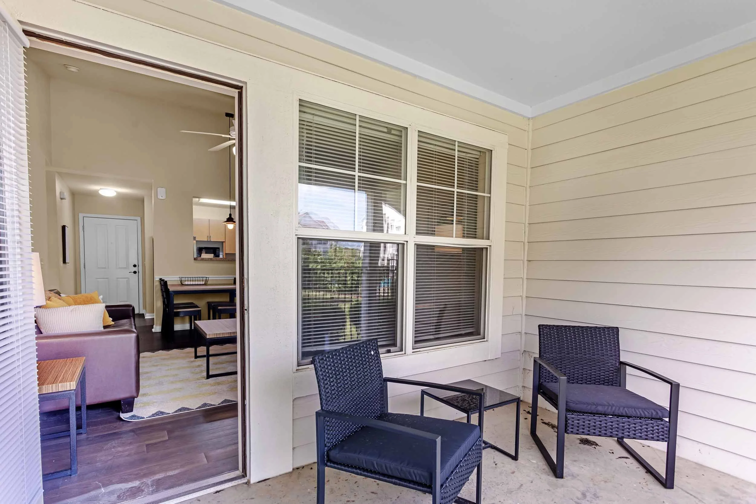 View of a covered outdoor patio area with black chairs, beige and yellow cushions, a small side table, and a sliding glass door leading into a living room with dining table and chairs, interior lighting, and beige walls.