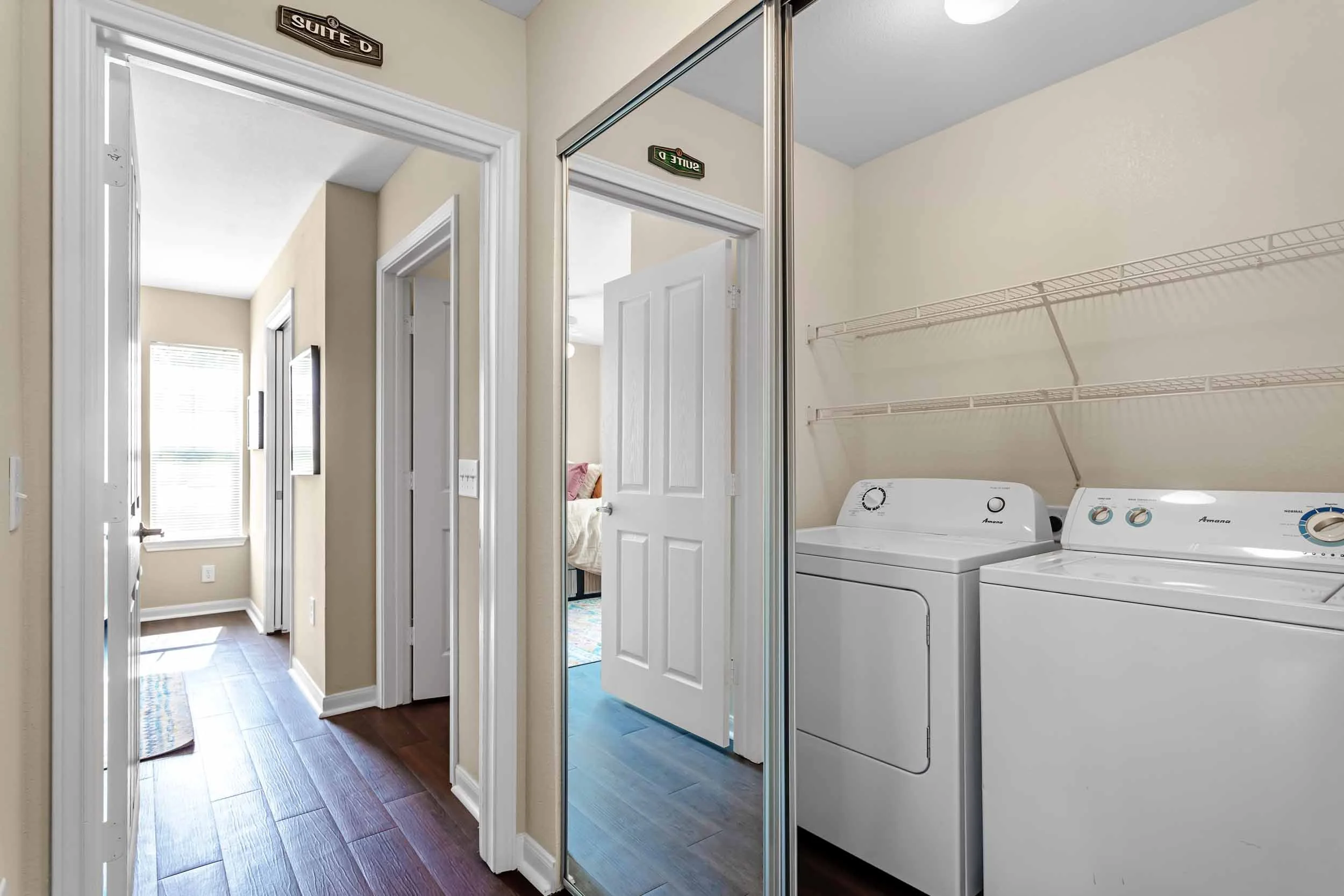 Inside a laundry room with a side-by-side washer and dryer, mirrored closet doors, and hallway leading to a bedroom with a bed and window