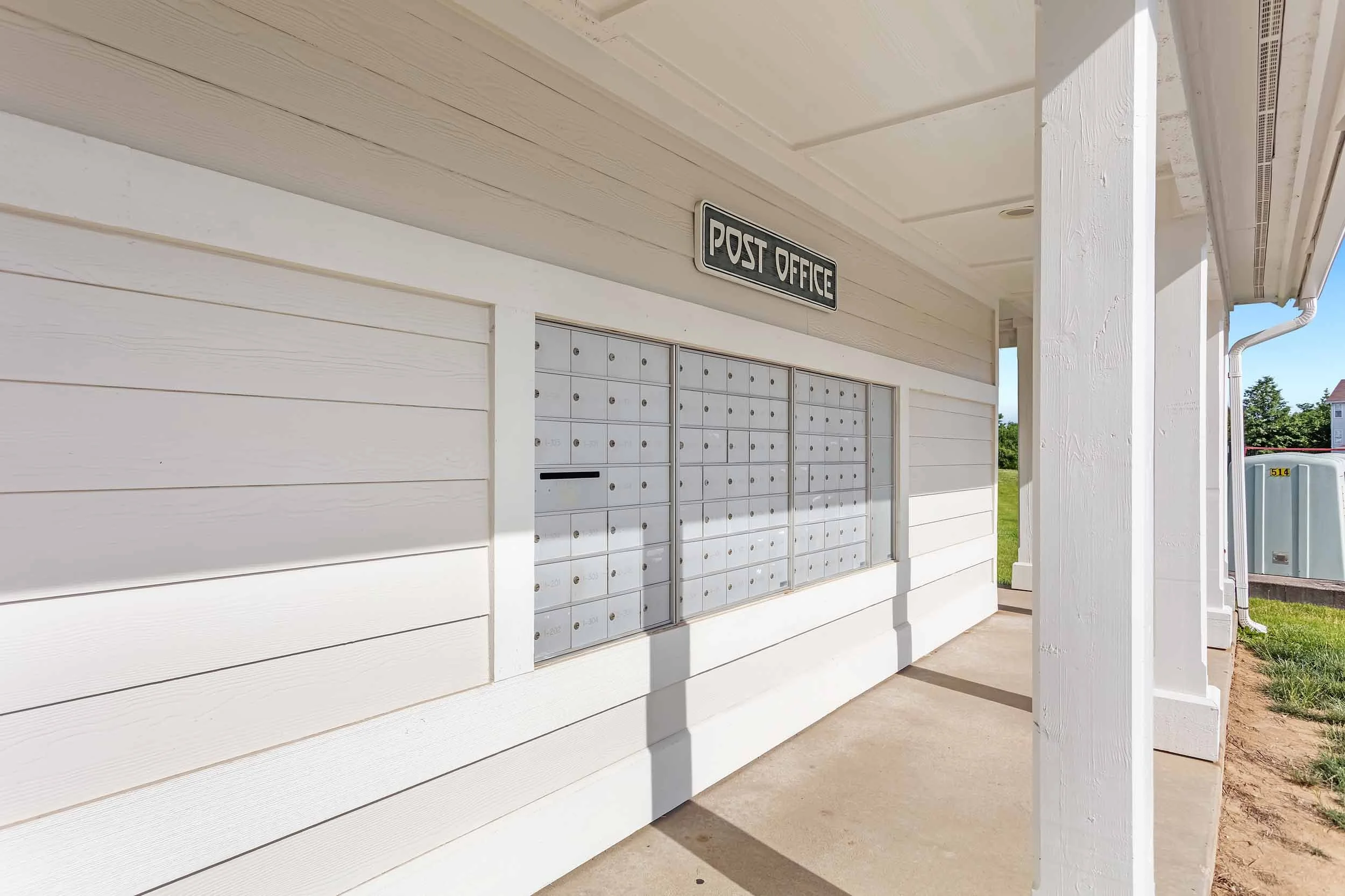 Multiple mailboxes on the wall, a sign that reads 'Post Office,' and a sidewalk in front, with clear skies and some greenery visible.