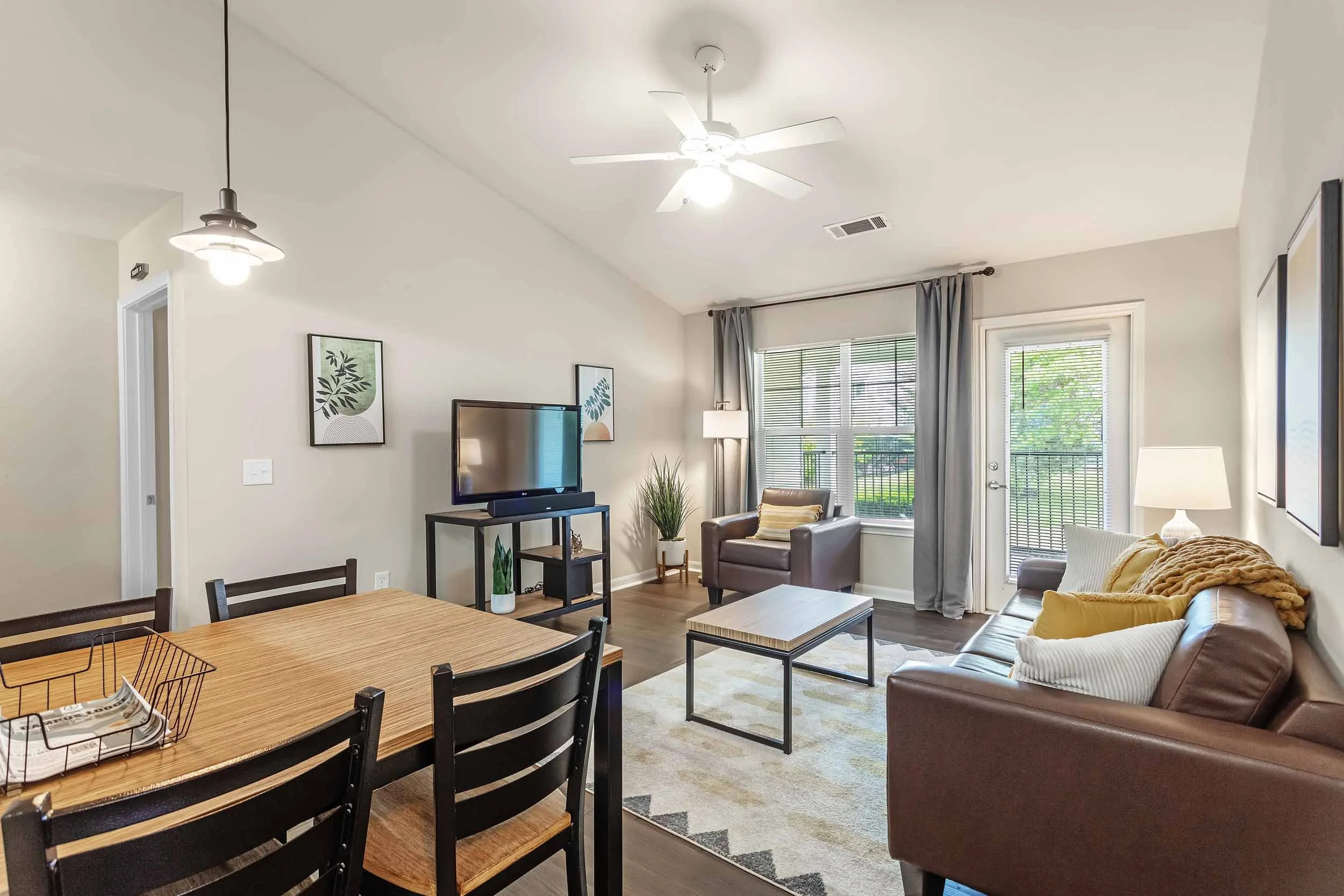 View of a cozy living room and dining area with a high vaulted ceiling, ceiling fan, large windows with blinds, a TV on a stand, a comfortable sofa, armchair, side table, floor lamp, and framed artwork on the walls in a well-lit space.