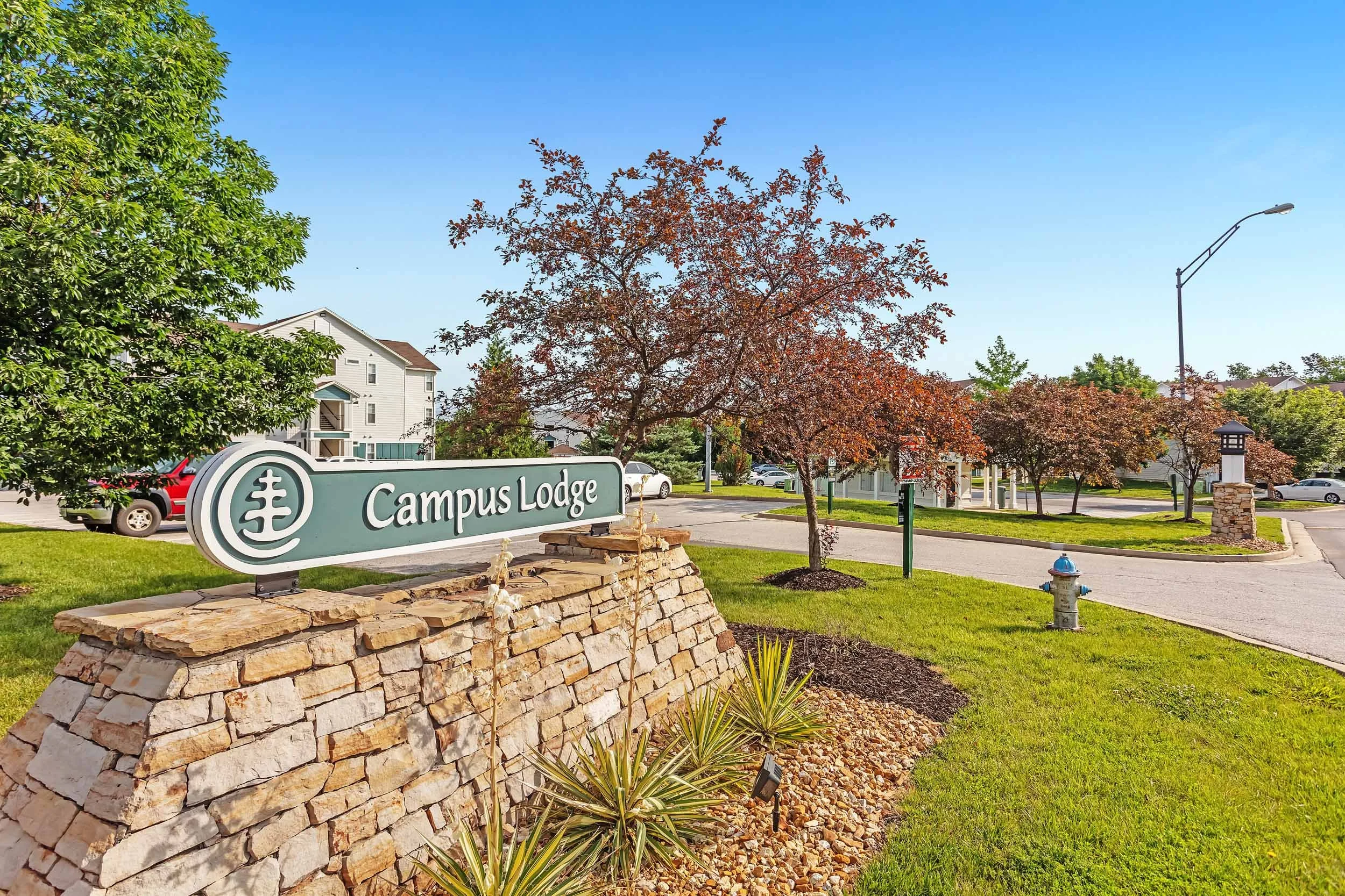 Sign for Campus Lodge in front of landscaped area with trees, grass, and small plants.