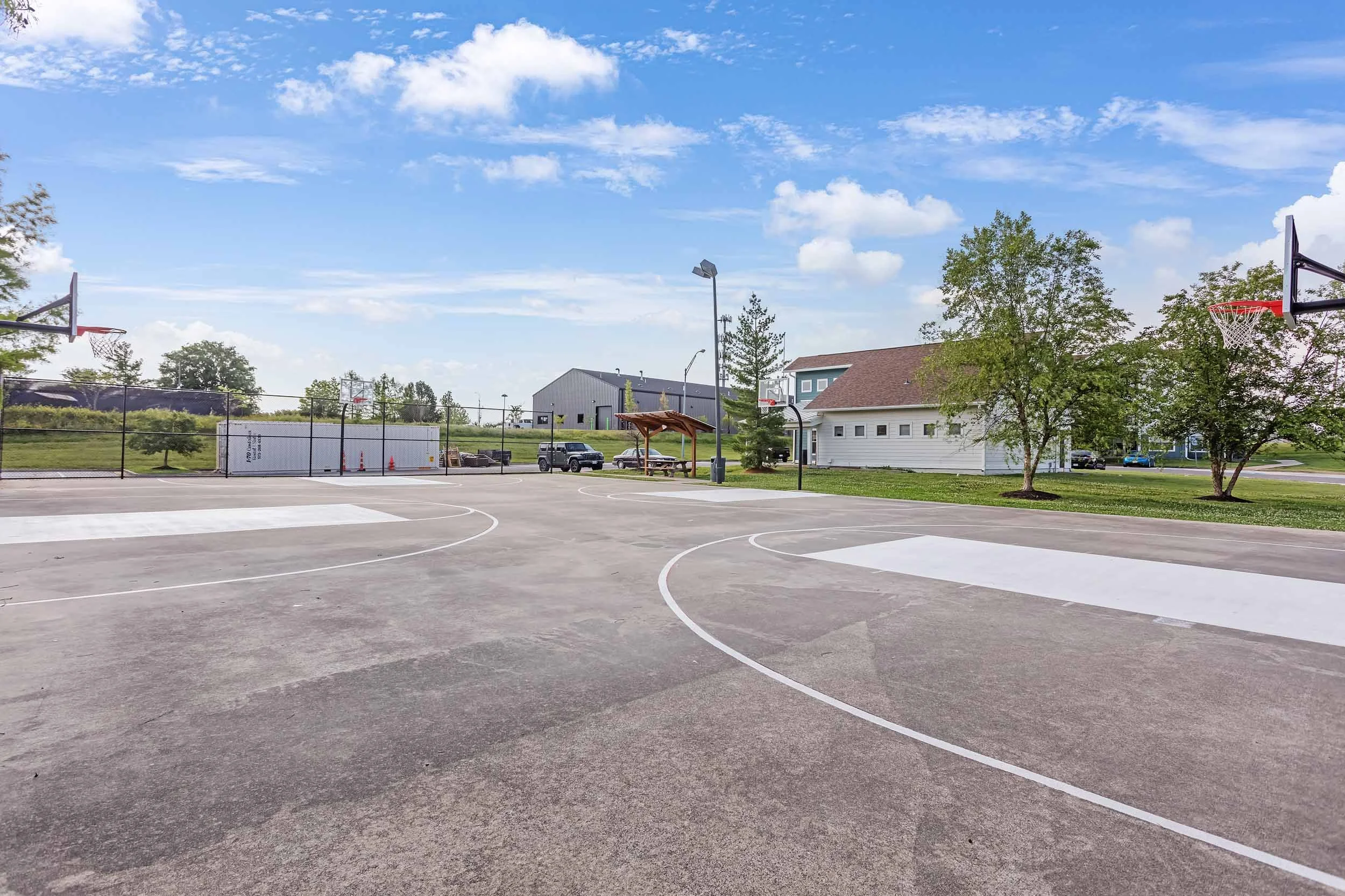 Empty outdoor basketball court with two basketball hoops, surrounded by trees and buildings on a sunny day with a blue sky and scattered clouds.