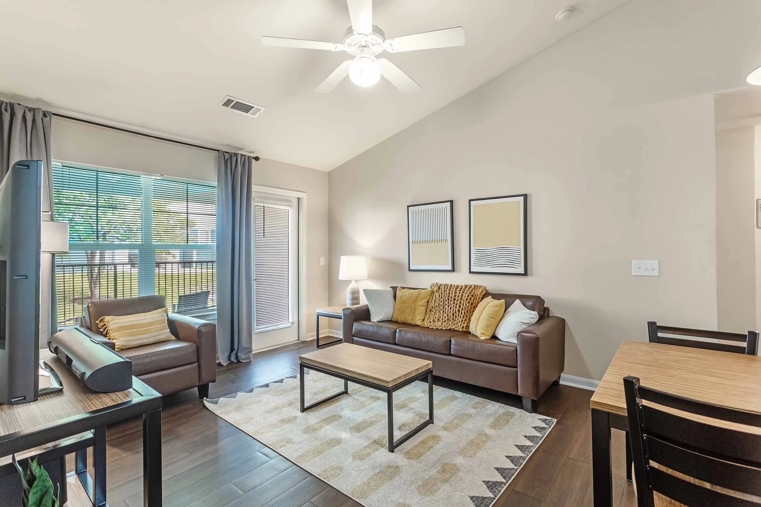 Living room with a ceiling fan, a large window with blinds and curtains, two brown sofas with pillows, a coffee table, a lamp, and framed artwork on the wall.