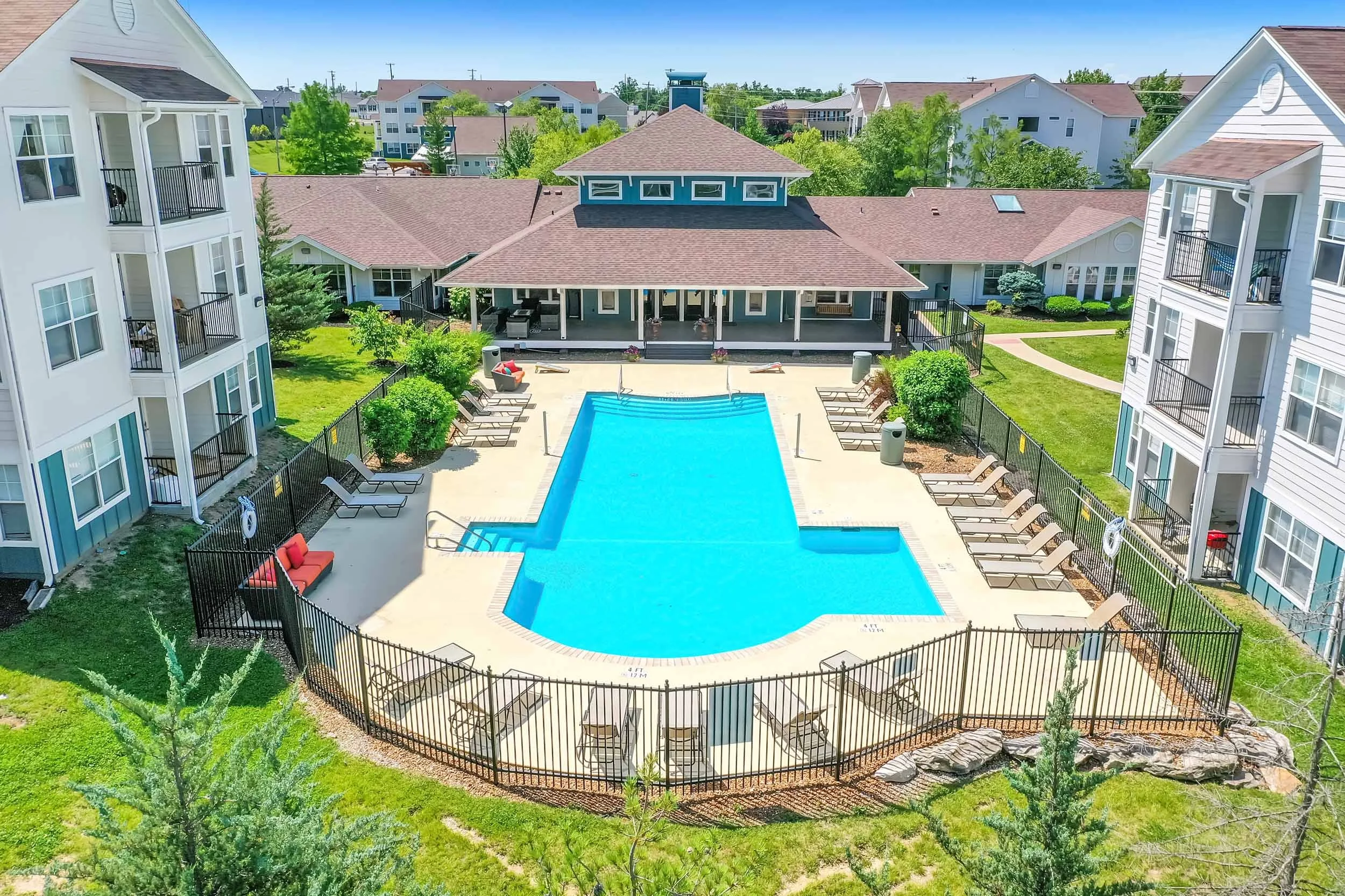 A large swimming pool surrounded by lounge chairs and a black metal fence in an apartment complex. There is a building with a porch and outdoor furniture behind the pool, with multi-story white residential buildings with balconies on either side and trees in the background.