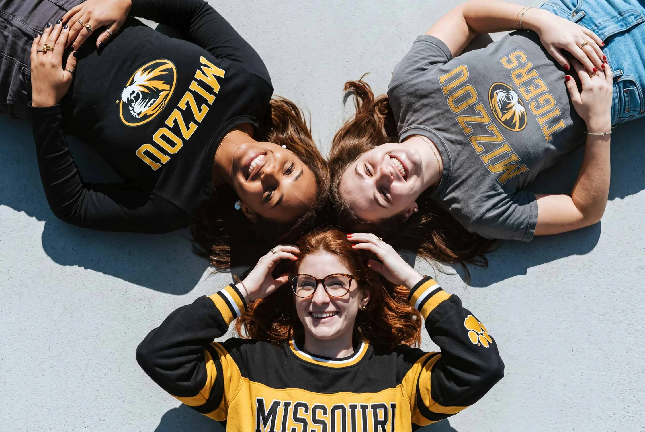 Four women lying on the ground in a row, smiling, wearing Missouri Tigers clothing, with their heads close together and hands on their chests, on a light-colored surface.