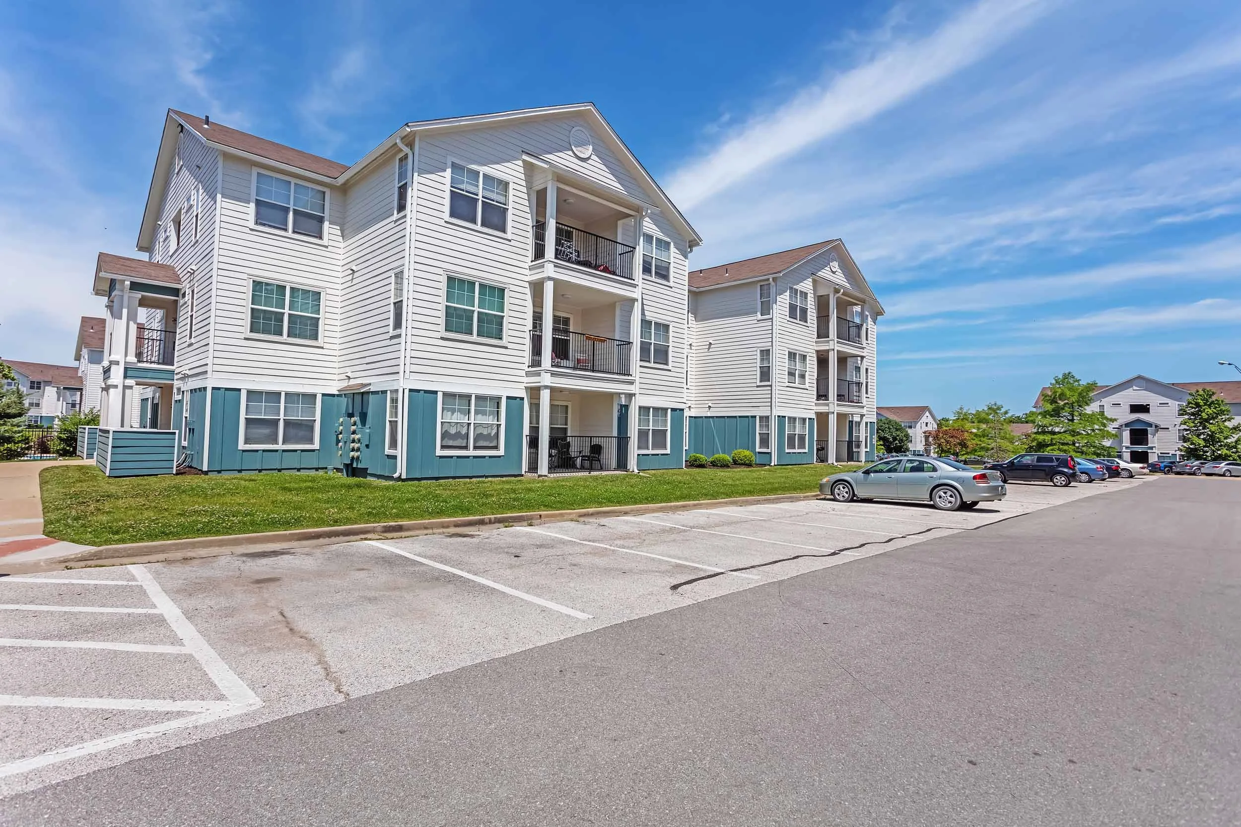 A large white multi-story apartment building with balconies and large windows, surrounded by a parking lot with a few parked cars, on a sunny day with a bright blue sky and some clouds.