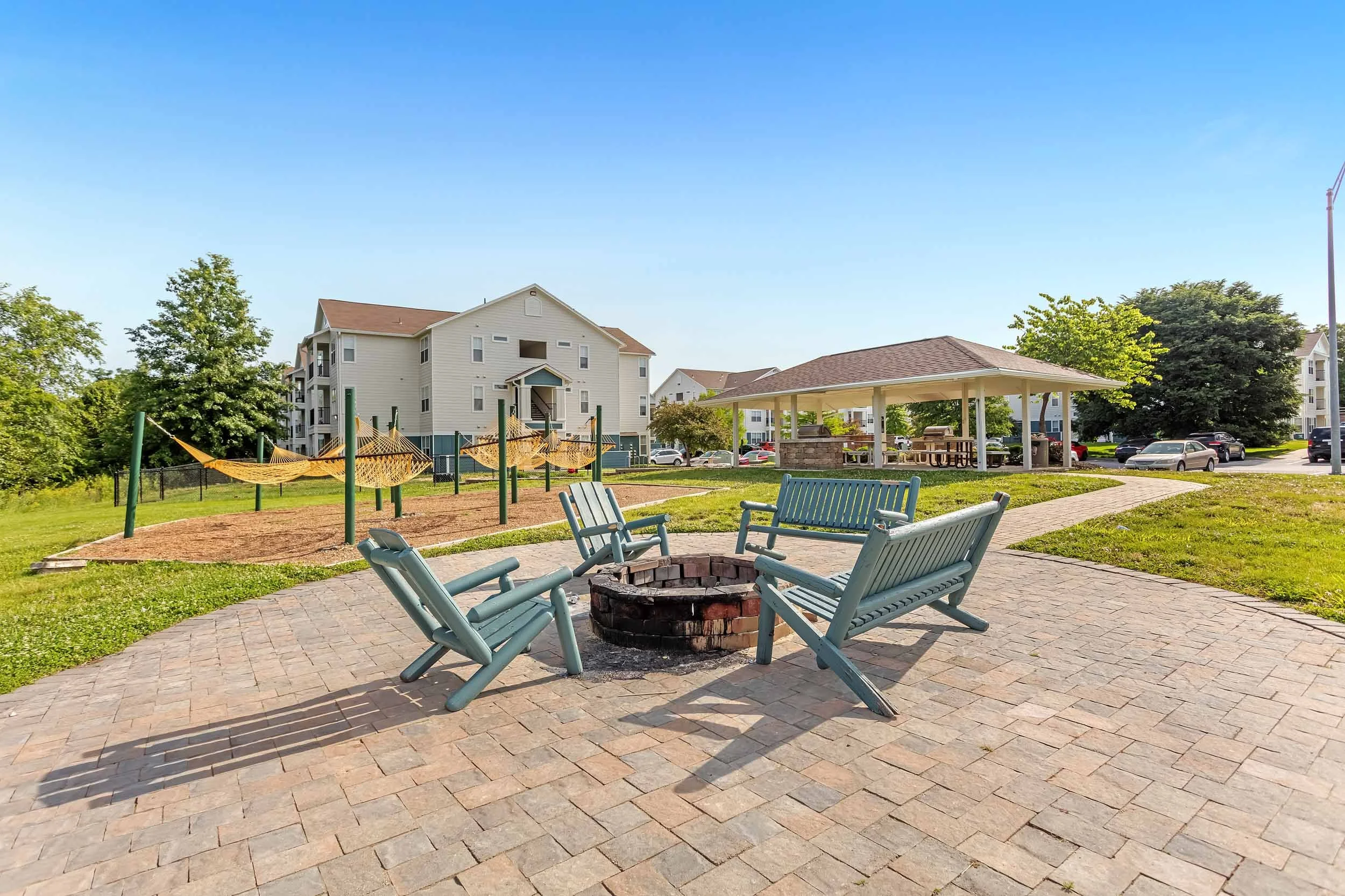 A park with a fire pit surrounded by four blue benches, a pavilion with picnic tables, a playground with hammock swings, and residential buildings in the background under a clear blue sky.