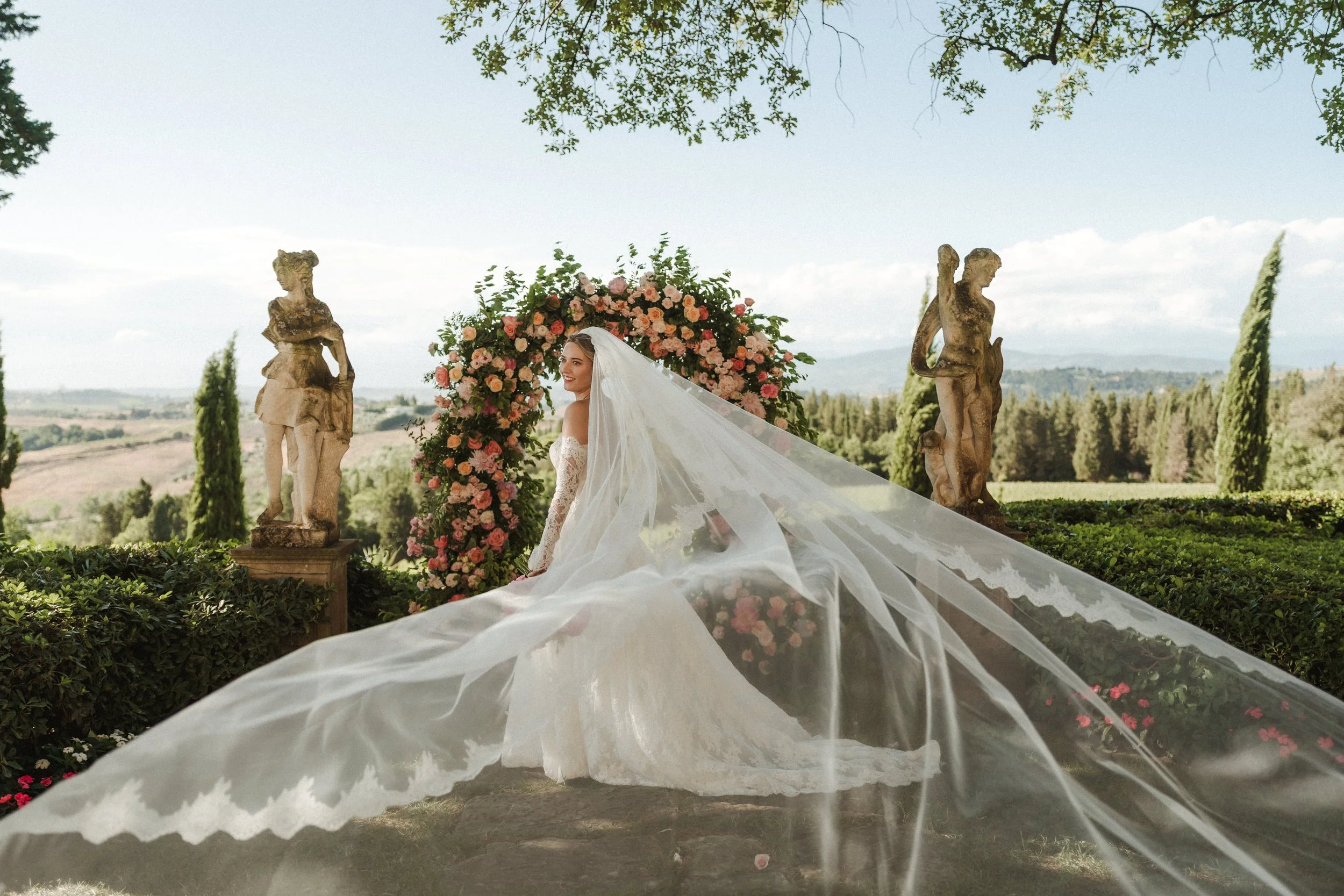 A bride in a wedding dress with a long train and veil standing outdoors near a floral arch and classical statues, with a scenic landscape in the background.