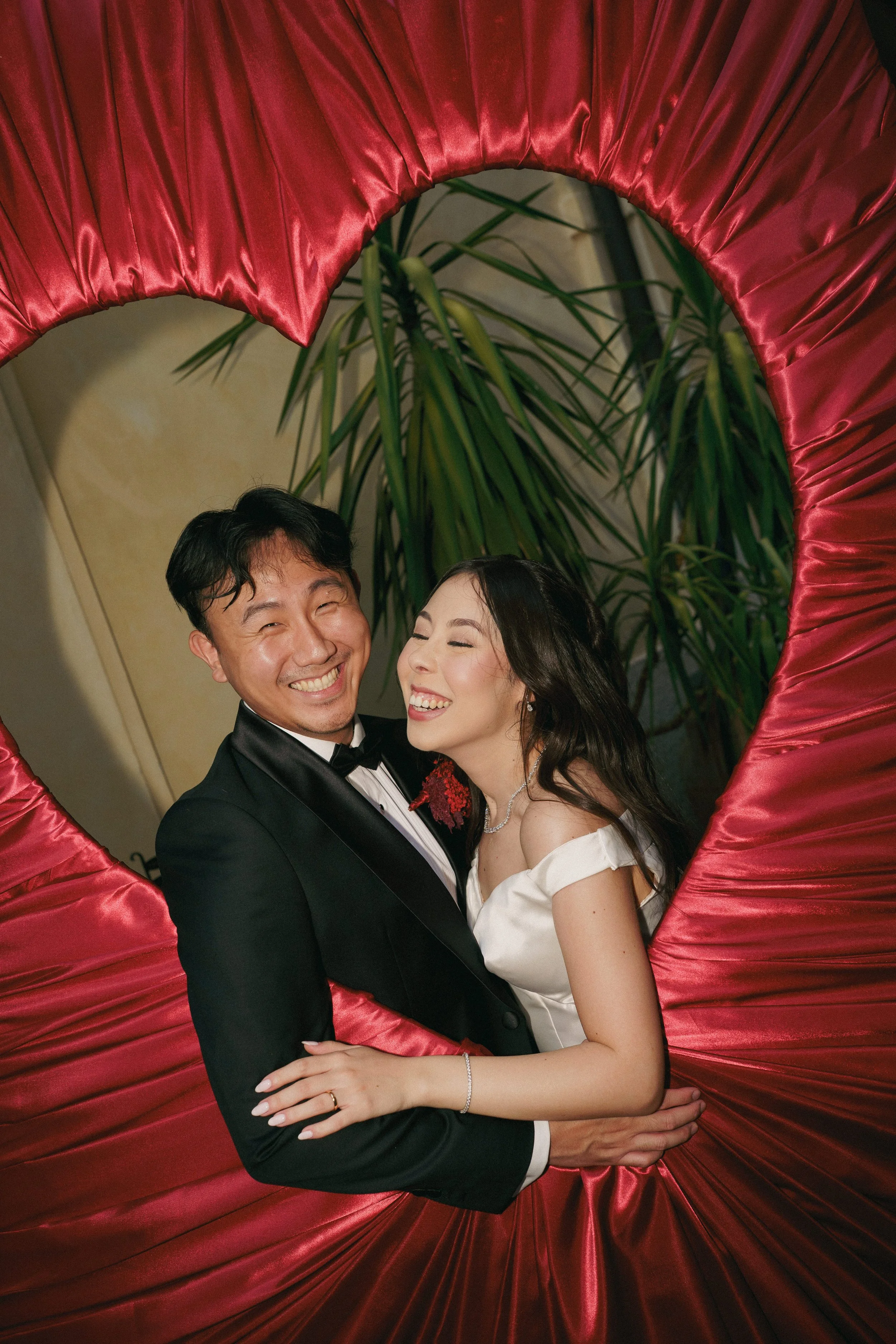 A happy couple dressed in formal attire embracing each other under a large, heart-shaped red satin decoration with lush green plants in the background.