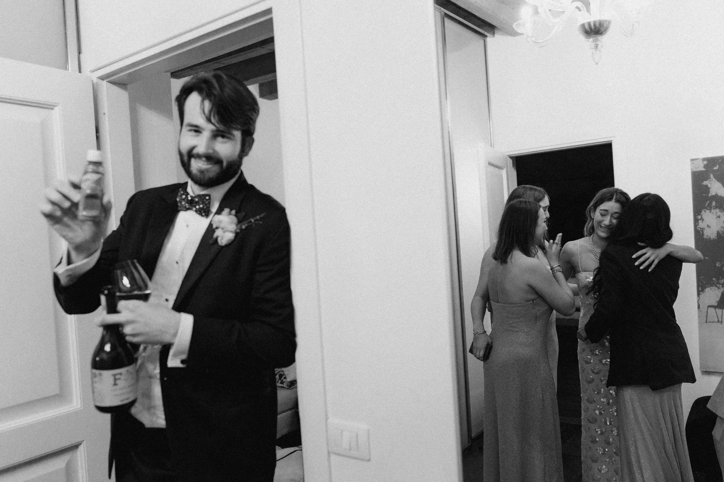 A man in a tuxedo with a bow tie holding a bottle of wine and a glass, smiling, at a social event. In the background, a group of women in formal dresses are hugging and chatting inside a well-lit room.