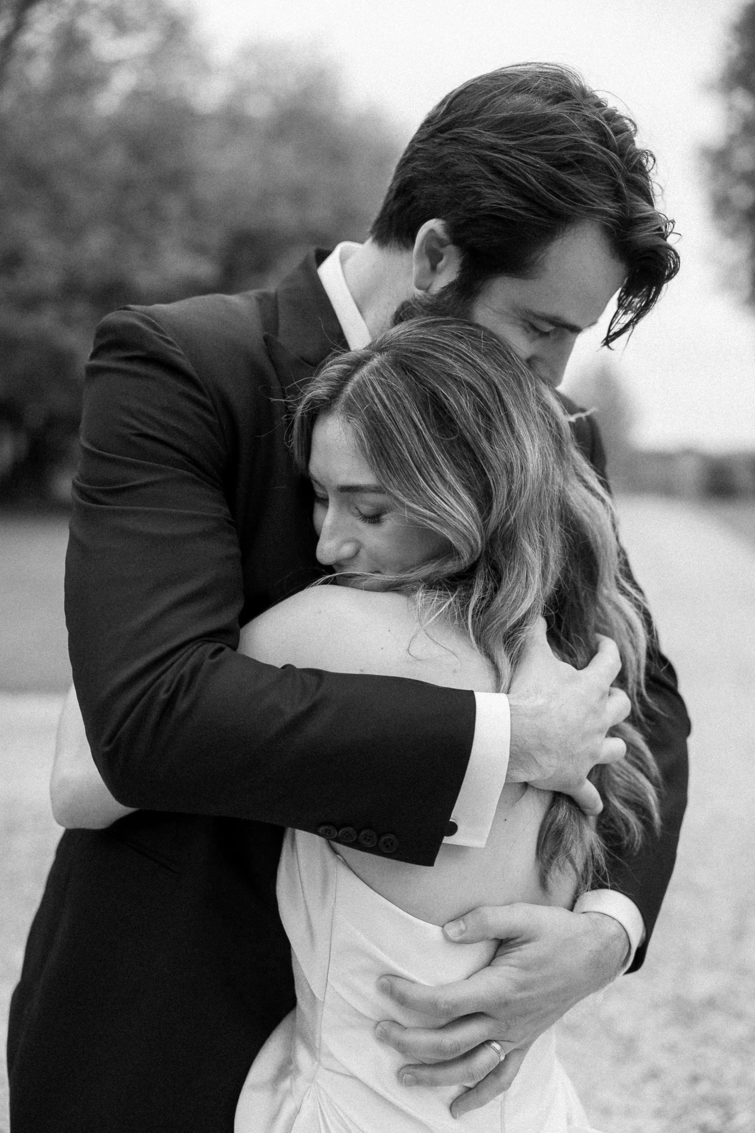 A black and white photo of a couple embracing, with the man wearing a suit and the woman in a wedding dress, outdoors with trees in the background.
