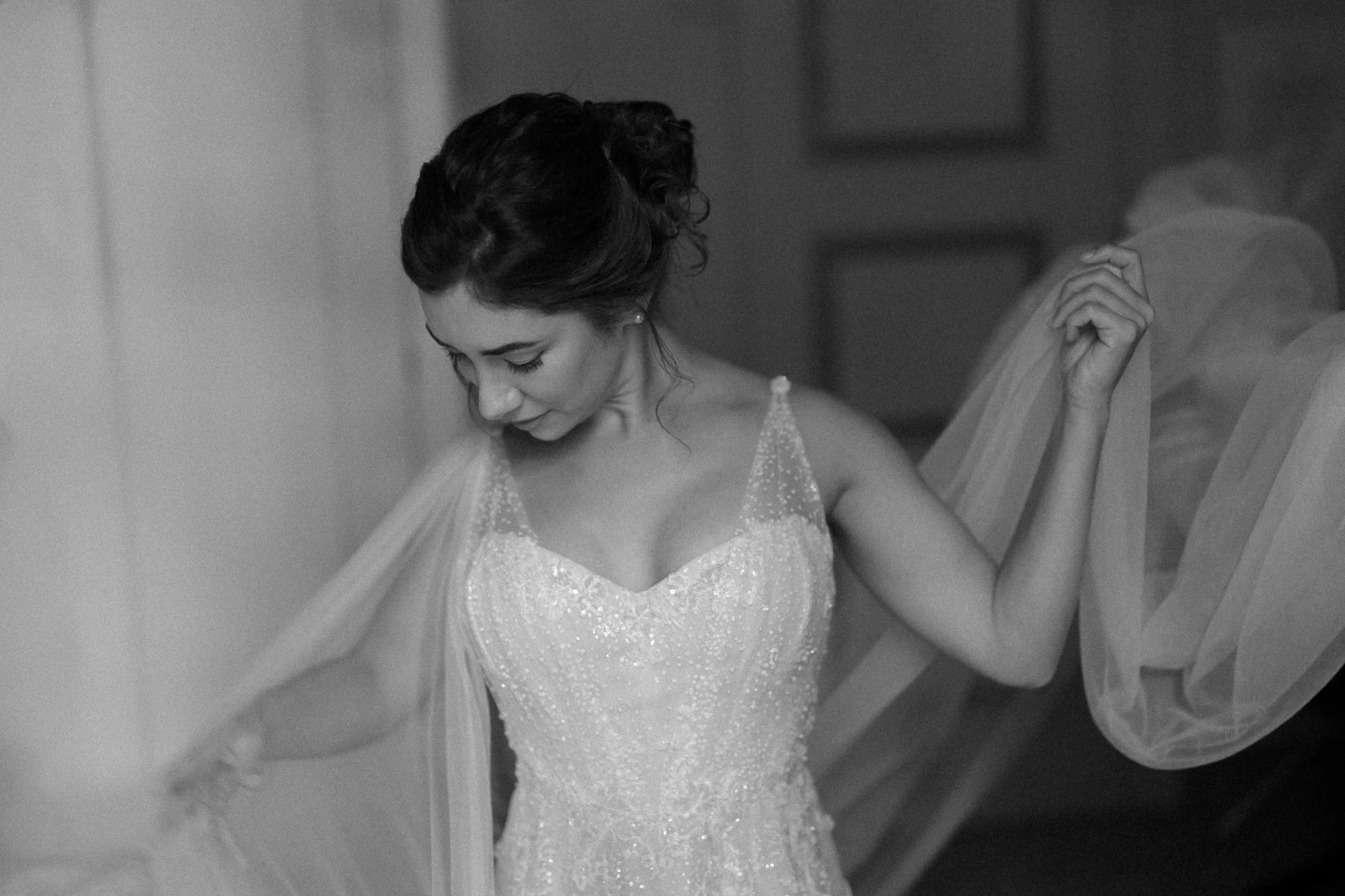 A woman in a wedding gown with a beaded bodice and sheer tulle sleeves, with dark hair styled in an updo, looking down while holding the edge of her veil.