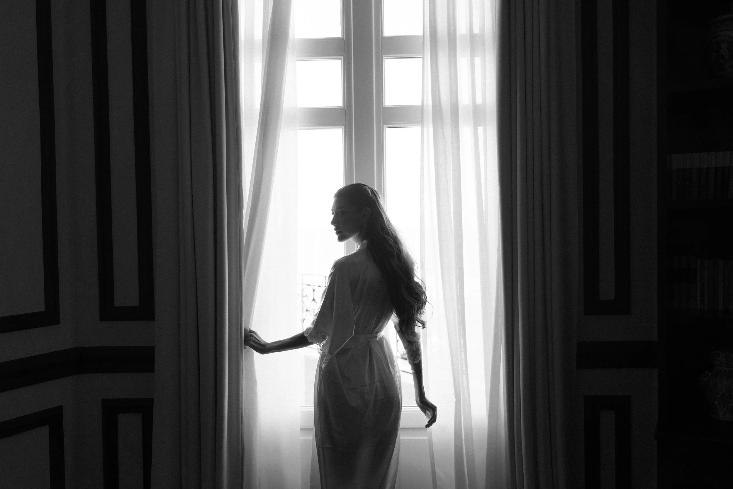 A silhouette of a woman in a white dress standing near a large window with sheer curtains, with sunlight streaming in.