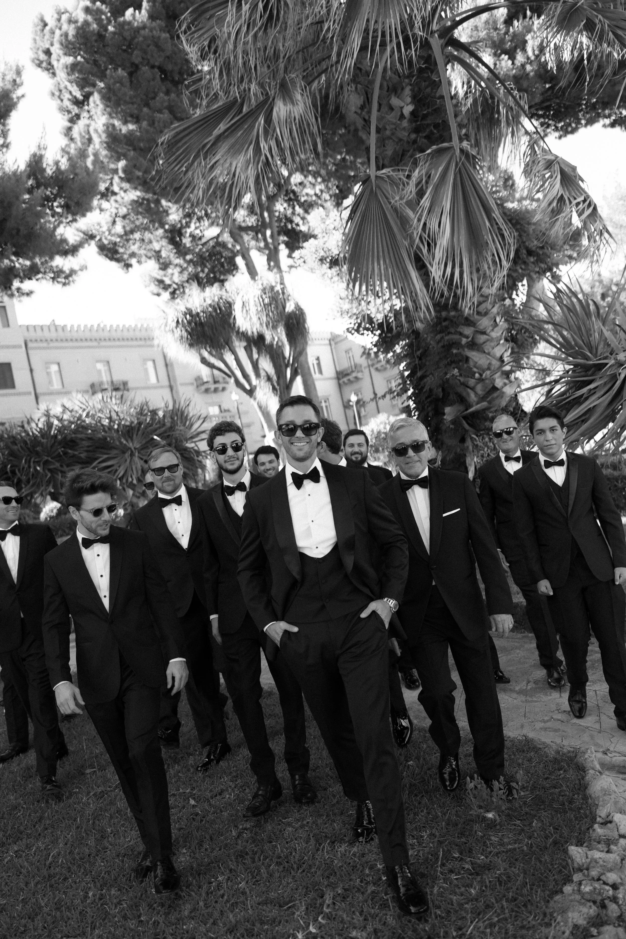 A group of men in tuxedos and sunglasses walking outdoors under trees.