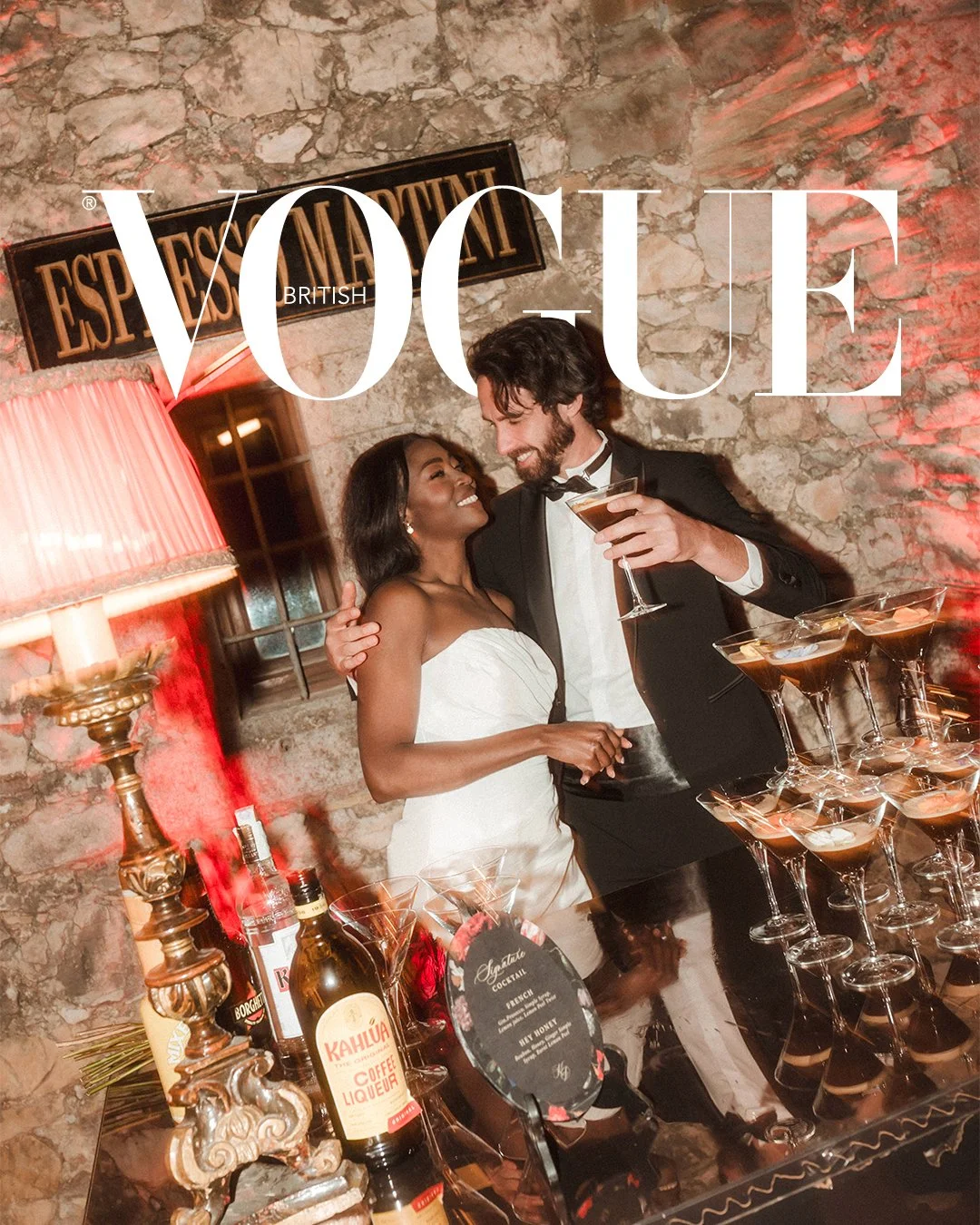 Two people, a woman in a white strapless dress and a man in a black tuxedo, are smiling and celebrating with cocktails at a bar. The setting has a rustic stone wall and a sign that reads "Esparit". There are bottles of alcohol and a tiered display of martini glasses on the table.