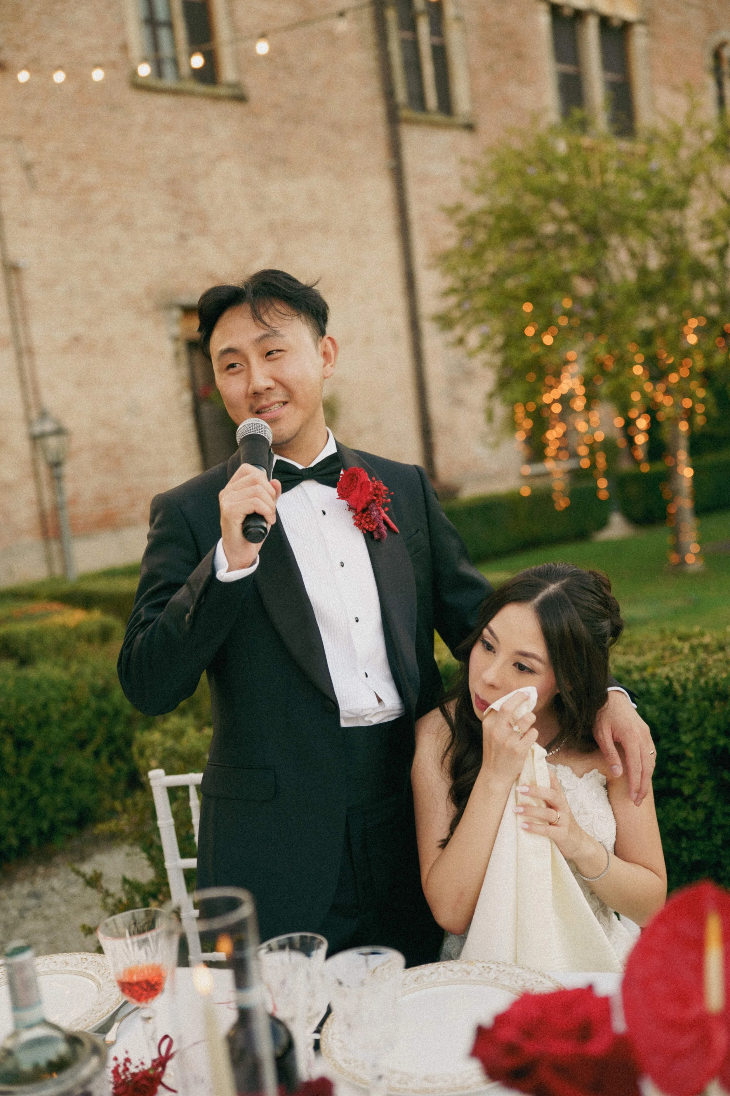 A man in a black tuxedo and bow tie holding a microphone, standing next to a woman in a wedding dress at an outdoor wedding reception. The man is smiling and the woman appears emotional, wiping her face with a tissue. The background features a brick 