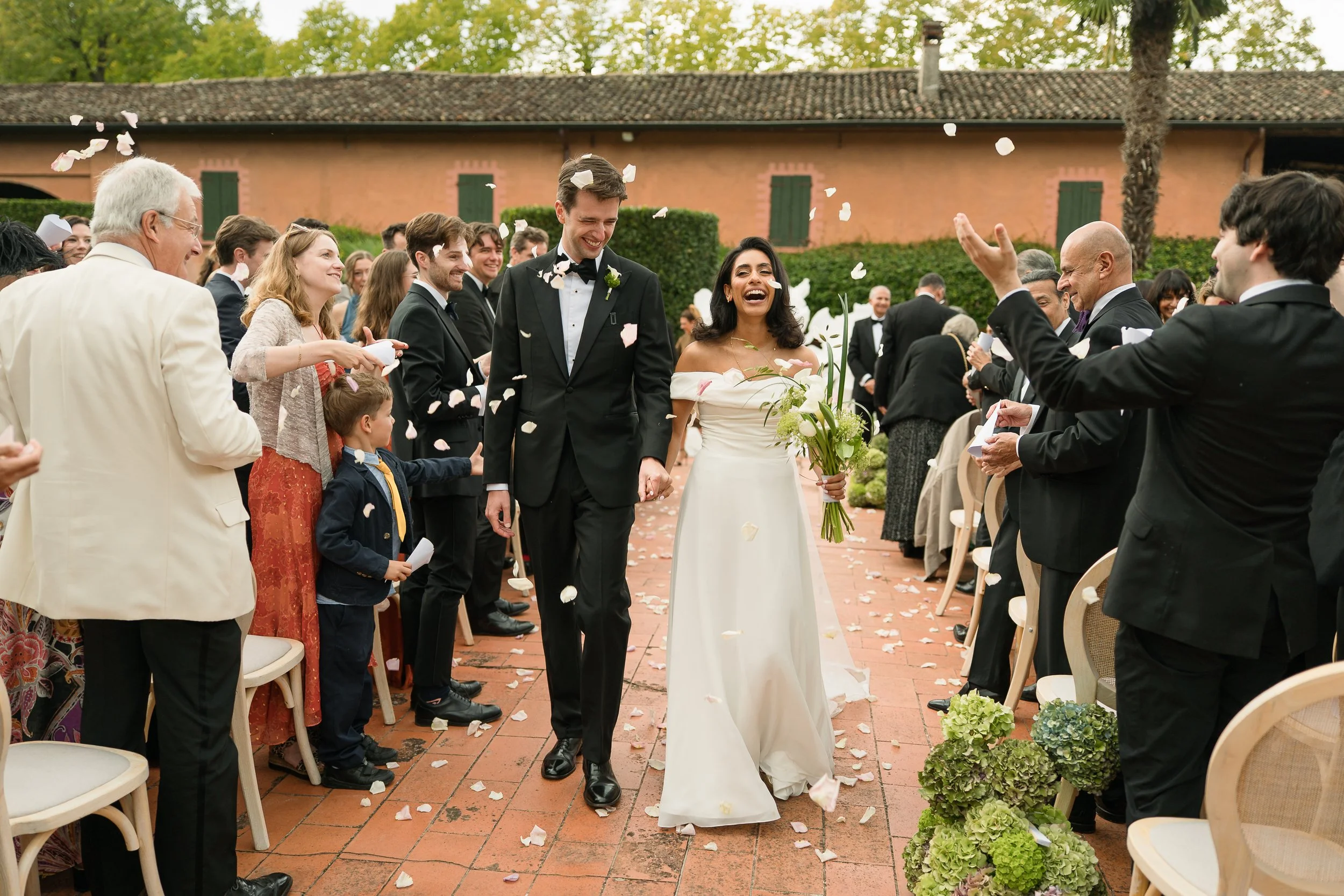 Bride and groom walking down the aisle during their wedding ceremony, surrounded by guests throwing flower petals and smiling. The bride is in a white off-shoulder gown holding a bouquet, while the groom is in a black tuxedo with a boutonniere. The o