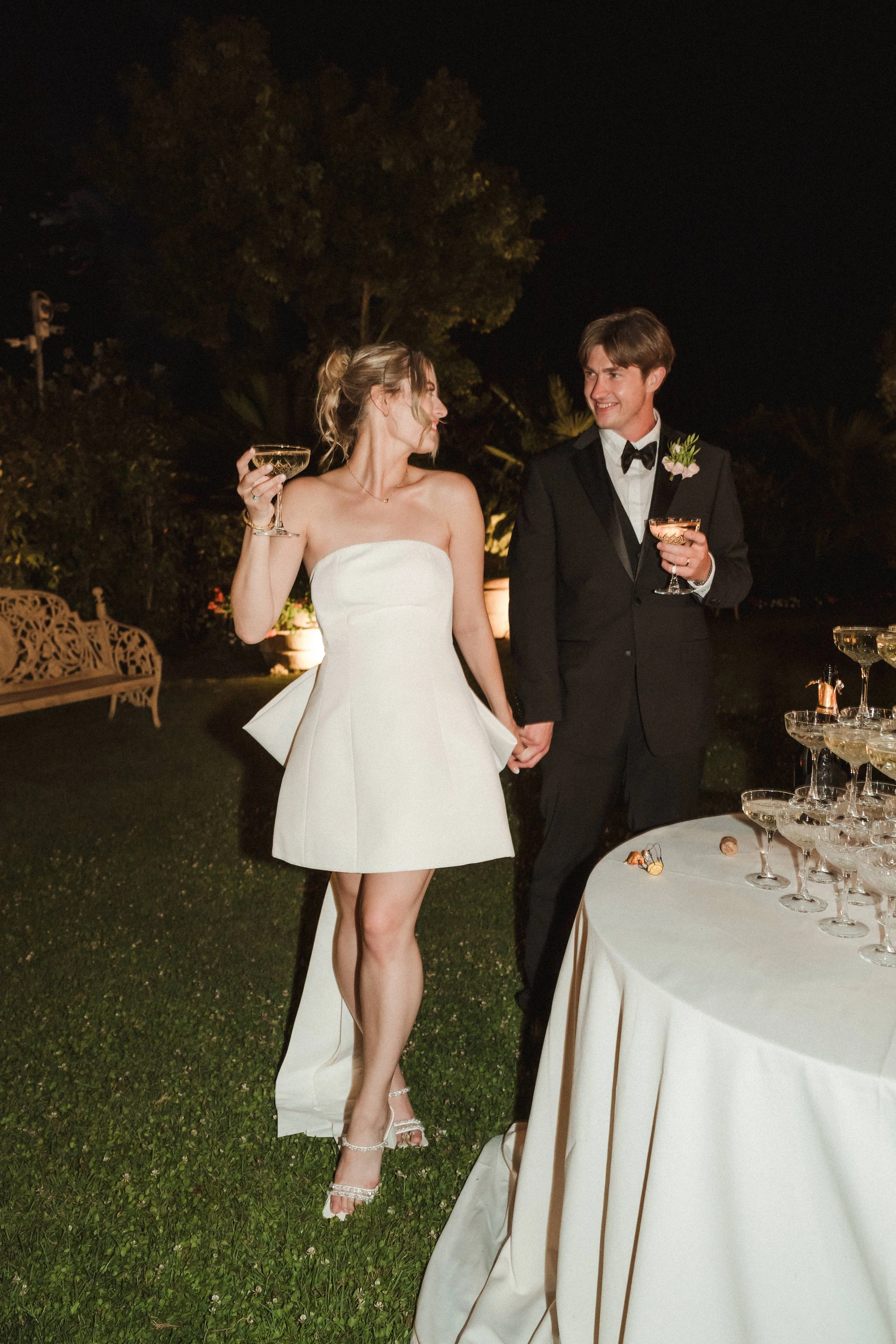 A bride and groom stand outdoors at night, holding glasses of champagne, smiling and looking at each other, with a table of champagne glasses in front of them.
