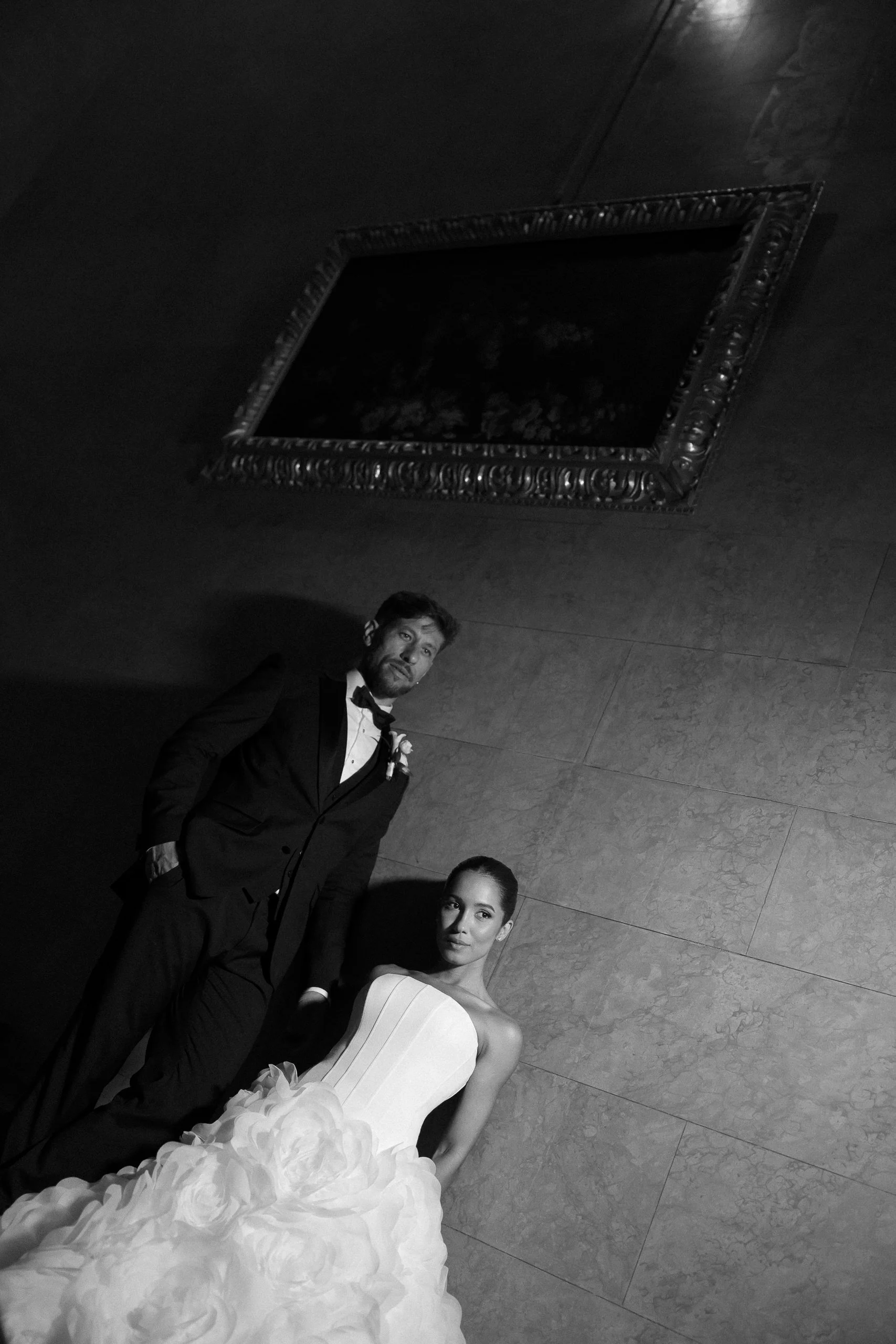 Black and white photograph of three people dressed formally, with a wall in the background, a decorative mirror on the ceiling, and a woman in an elegant strapless gown with a large floral detail at the bottom.