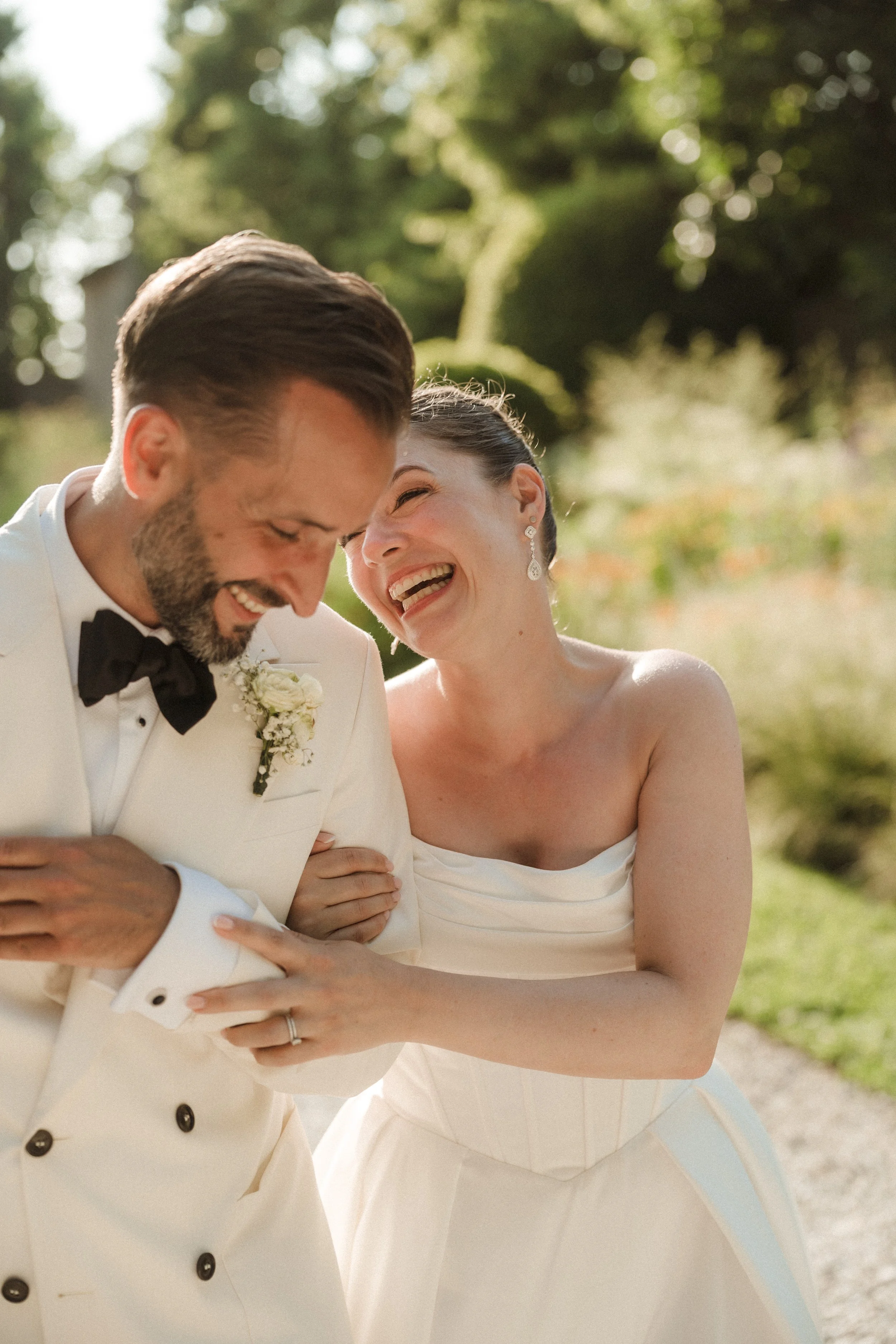 A joyful bride and groom laughing together outdoors, with the groom in a white tuxedo and the bride in a strapless white wedding dress, surrounded by greenery.