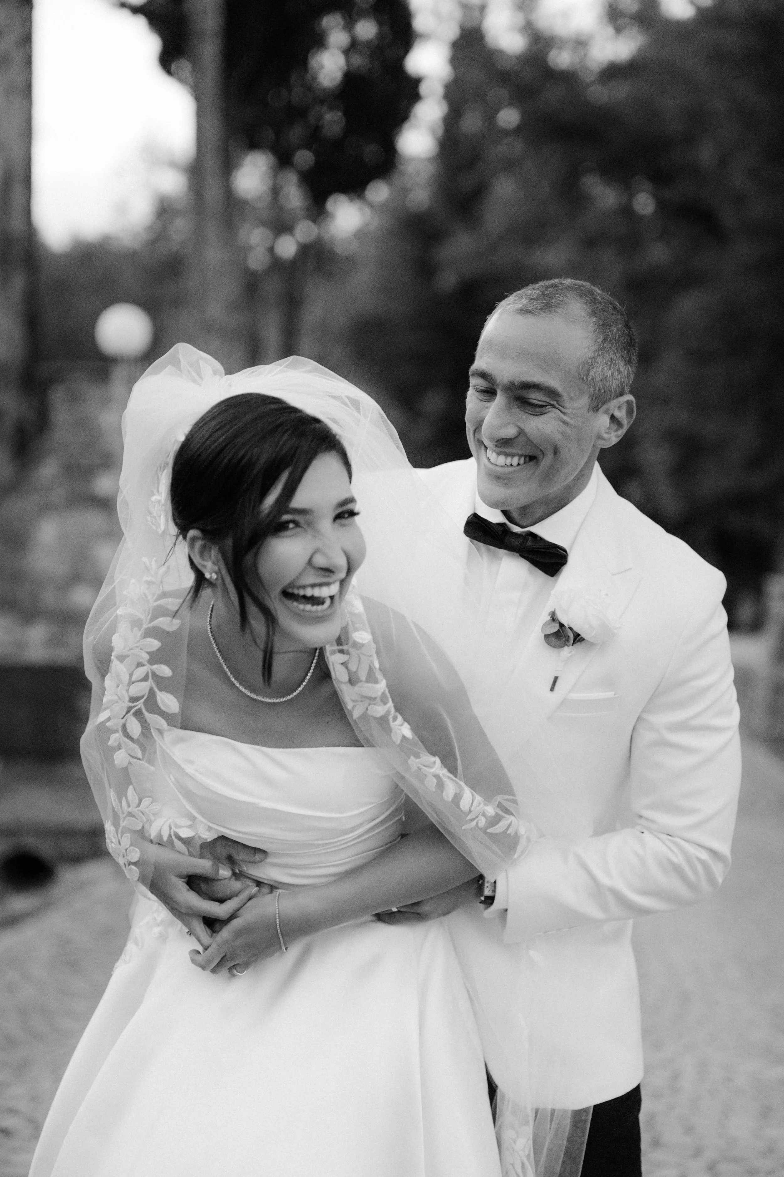 A black and white photo of a happy bride and groom. The bride wears a wedding dress and veil, and the groom in a white tuxedo with a black bow tie. They are smiling and embracing outdoors with trees in the background.
