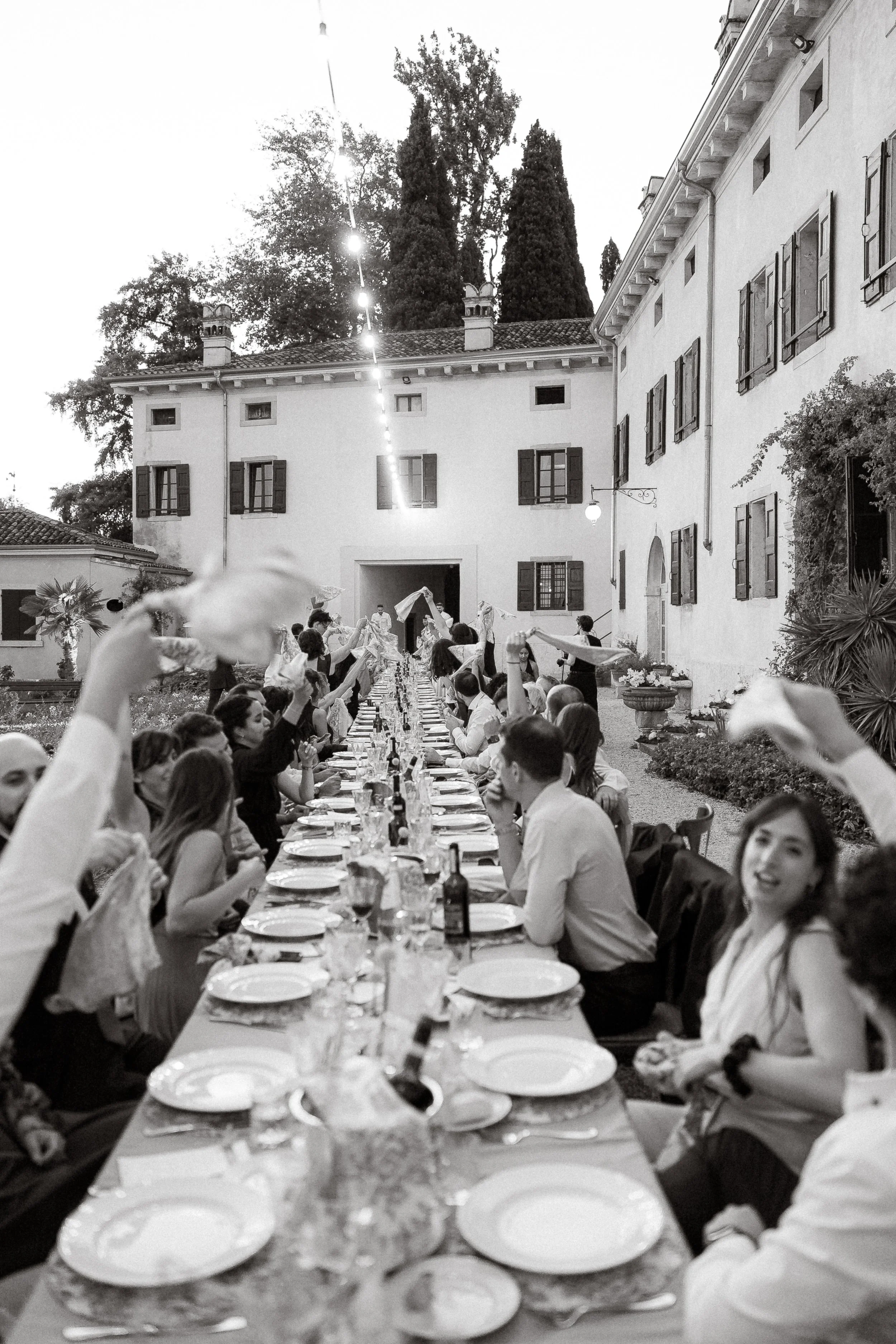 People seated at a long outdoor dining table with plates, glasses, and bottles, in front of a large white building with windows and shutters, under string lights and trees.