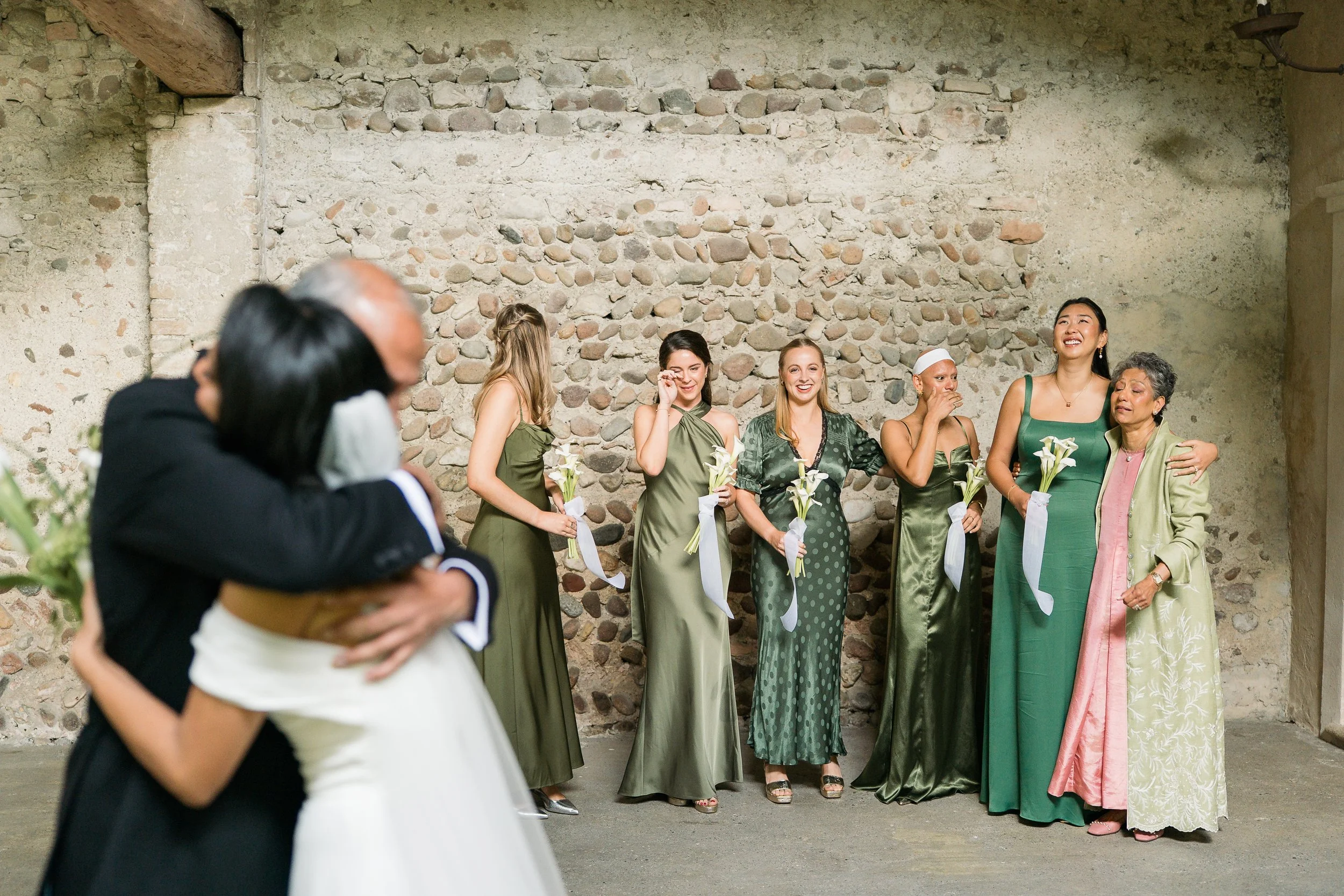 A group of women standing against a stone wall, holding white calla lilies, dressed in elegant green dresses, smiling and interacting during a celebration or event.