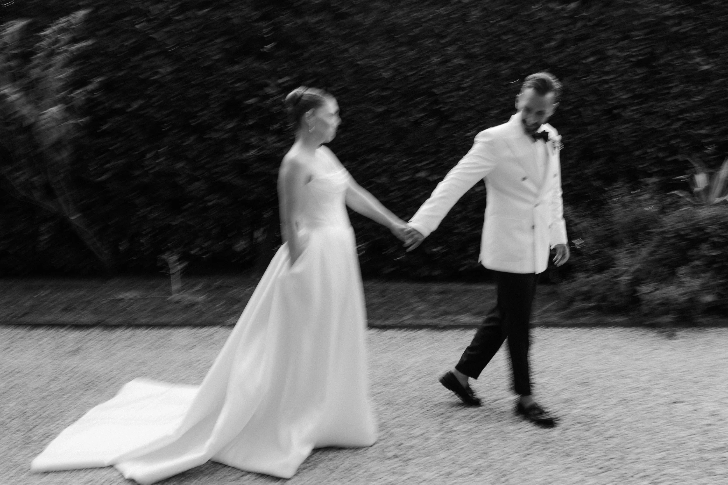 A bride and groom walking hand in hand outdoors, with the bride in a white wedding dress and the groom in a white tuxedo jacket with black bow tie, against a dark, blurry background.