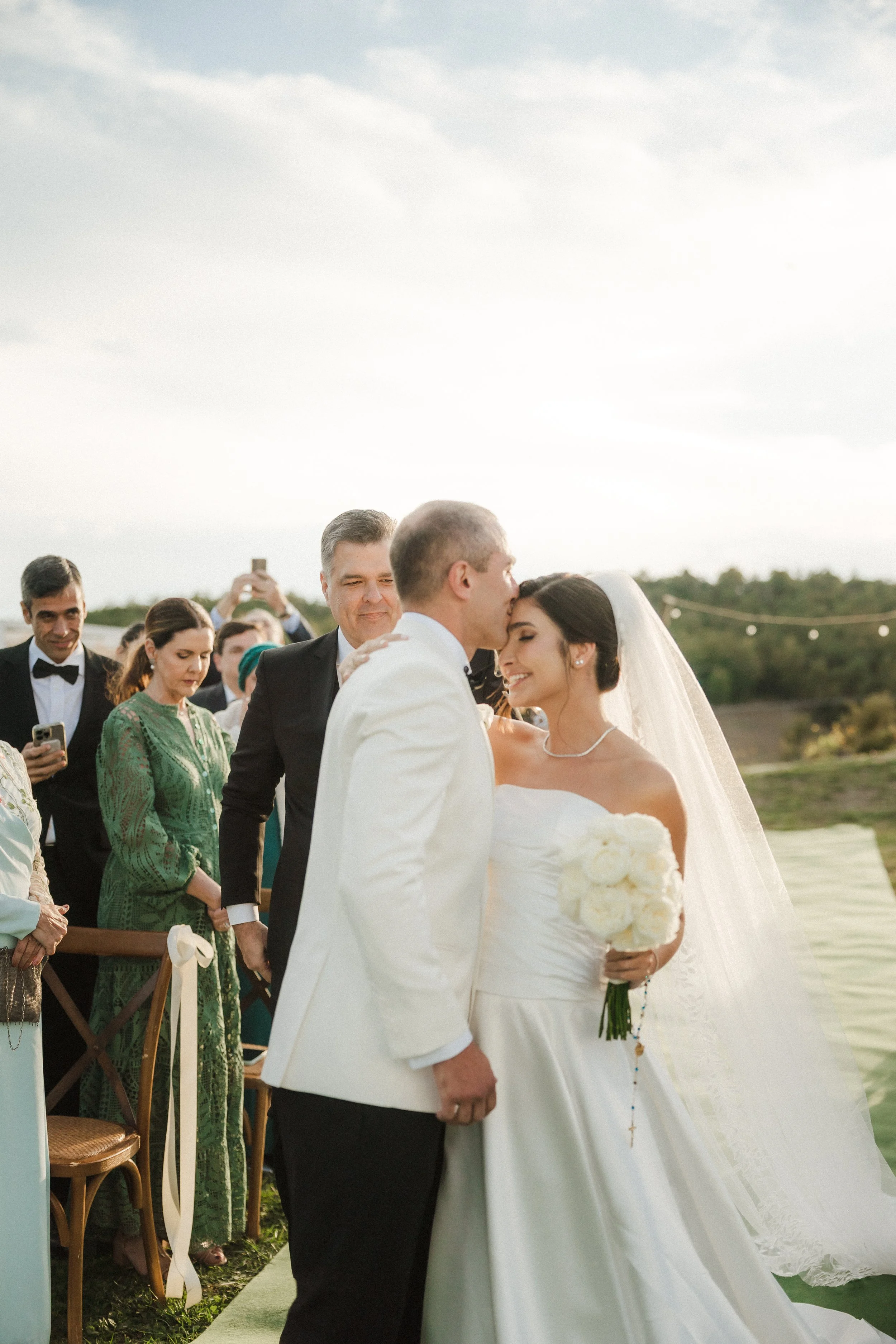A bride and groom kiss at their outdoor wedding ceremony, surrounded by guests in formal attire, with some taking photos, on a grassy field with a cloudy sky.