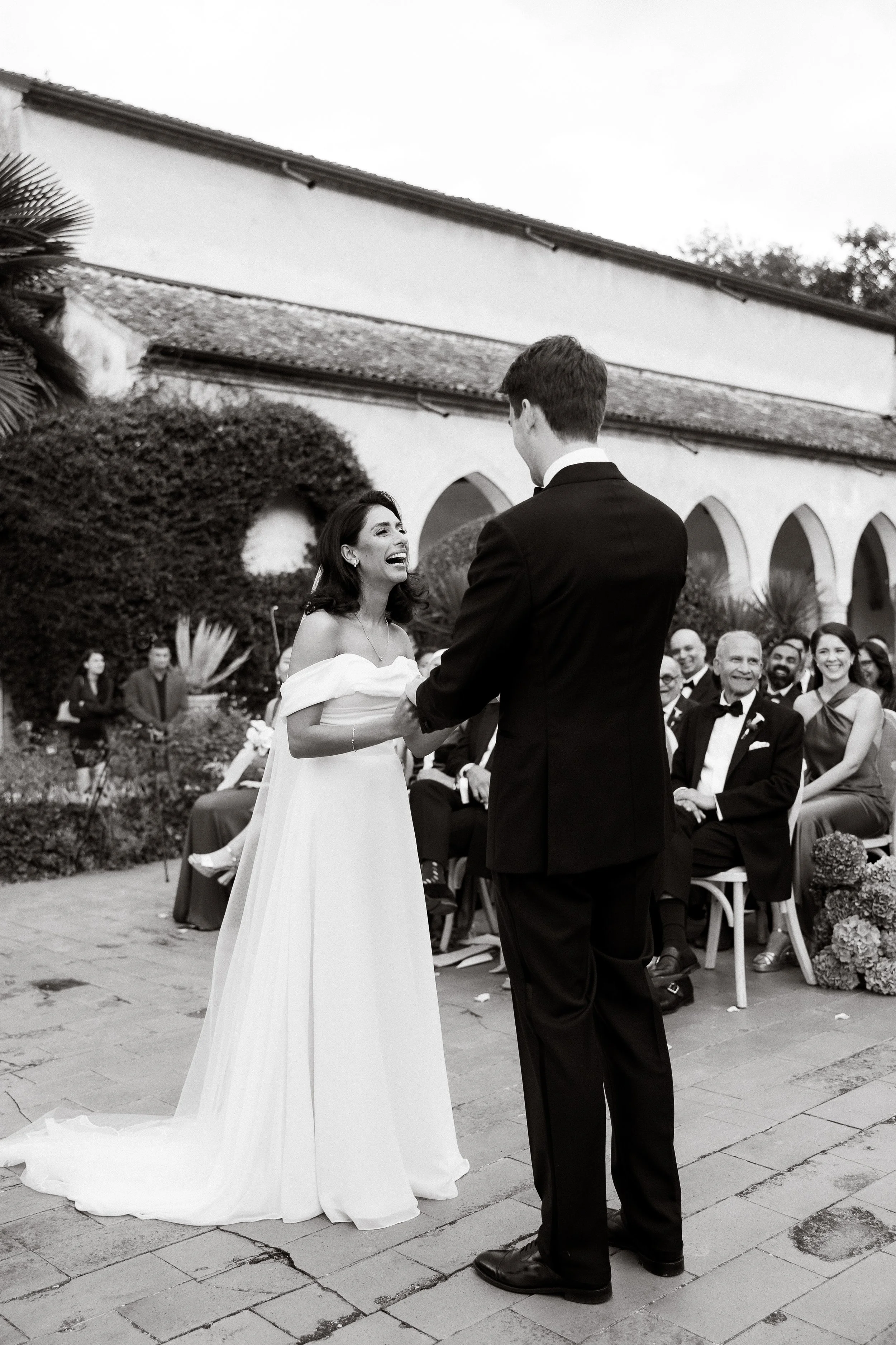 A bride and groom holding hands and exchanging vows during a wedding ceremony outdoors, with seated guests and a church building in the background.