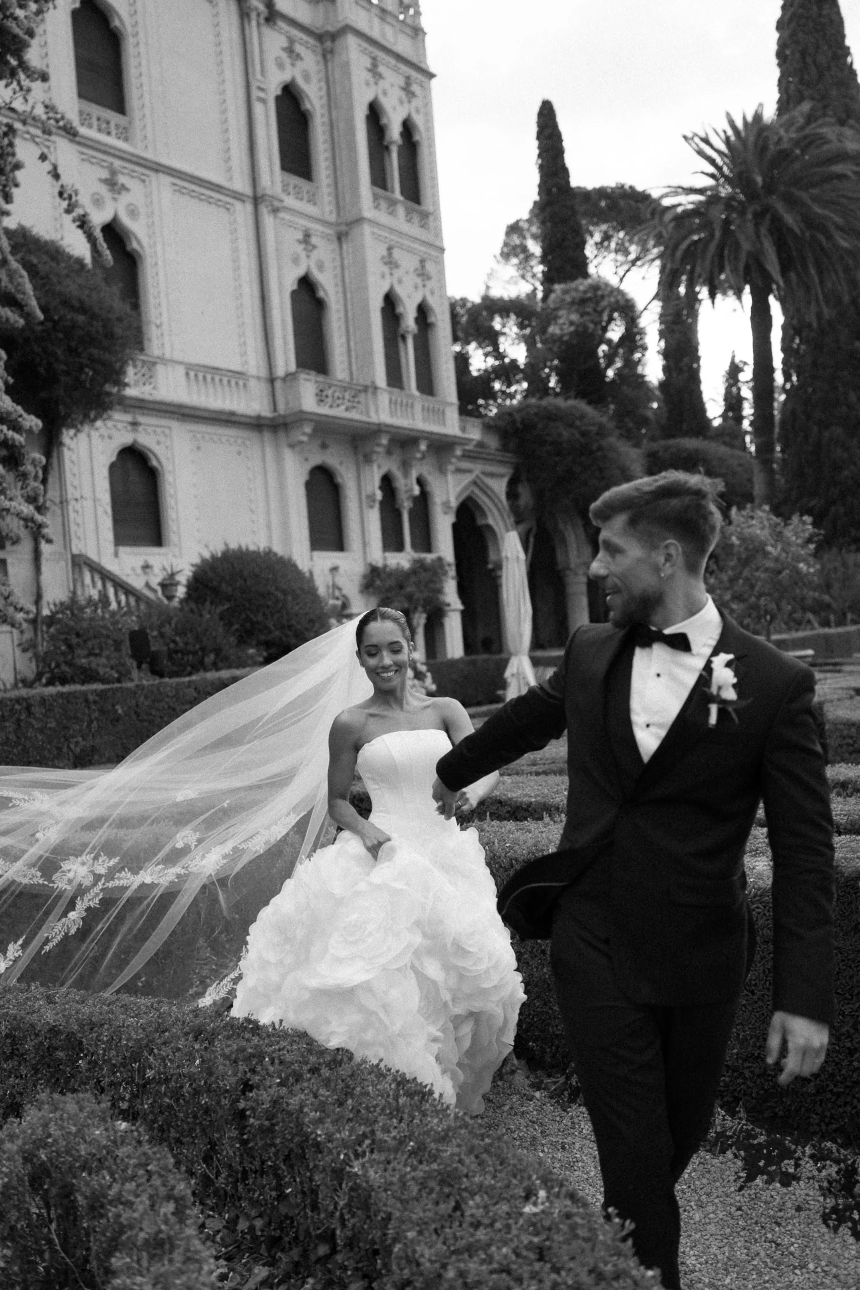A bride and groom are holding hands outdoors in front of a historic building with ornate architecture, surrounded by trees and shrubbery.