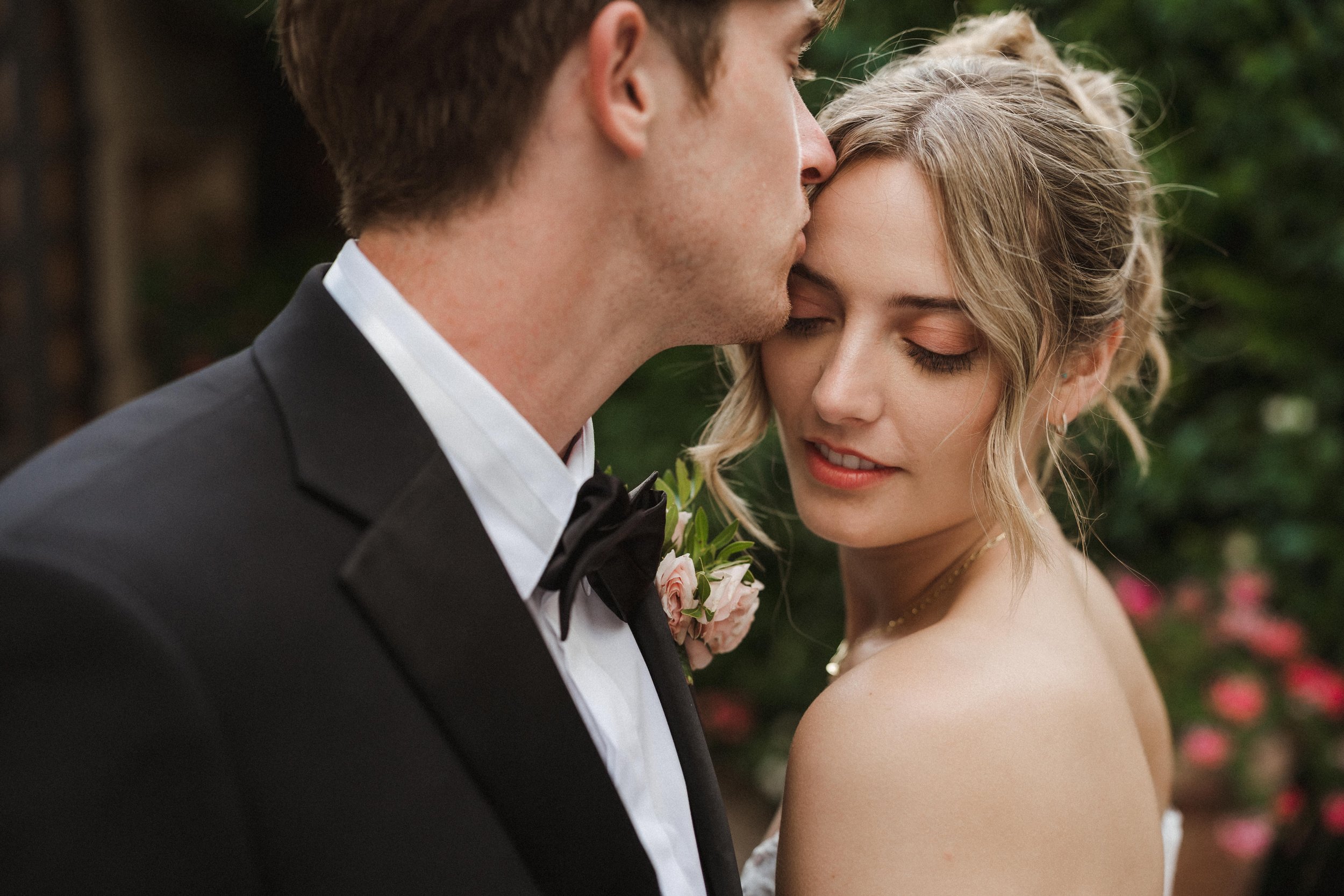A bride and groom share a close, intimate moment outdoors, with the groom kissing the bride's forehead and the bride gently closing her eyes, surrounded by greenery and pink flowers.