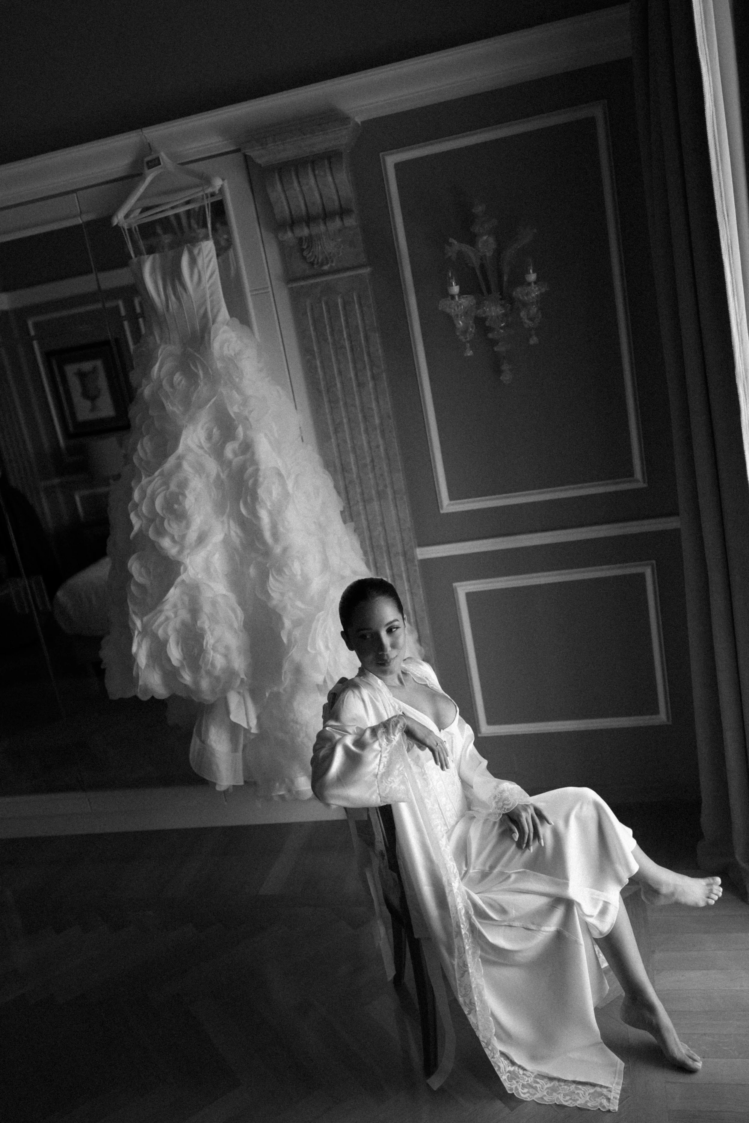 Black and white photo of a woman in a vintage dress sitting on a chair with her legs extended. She is in a room with decorated walls, a chandelier mirror, and a ruffled dress hanging from a rack. There is a young girl partially visible at the bottom 