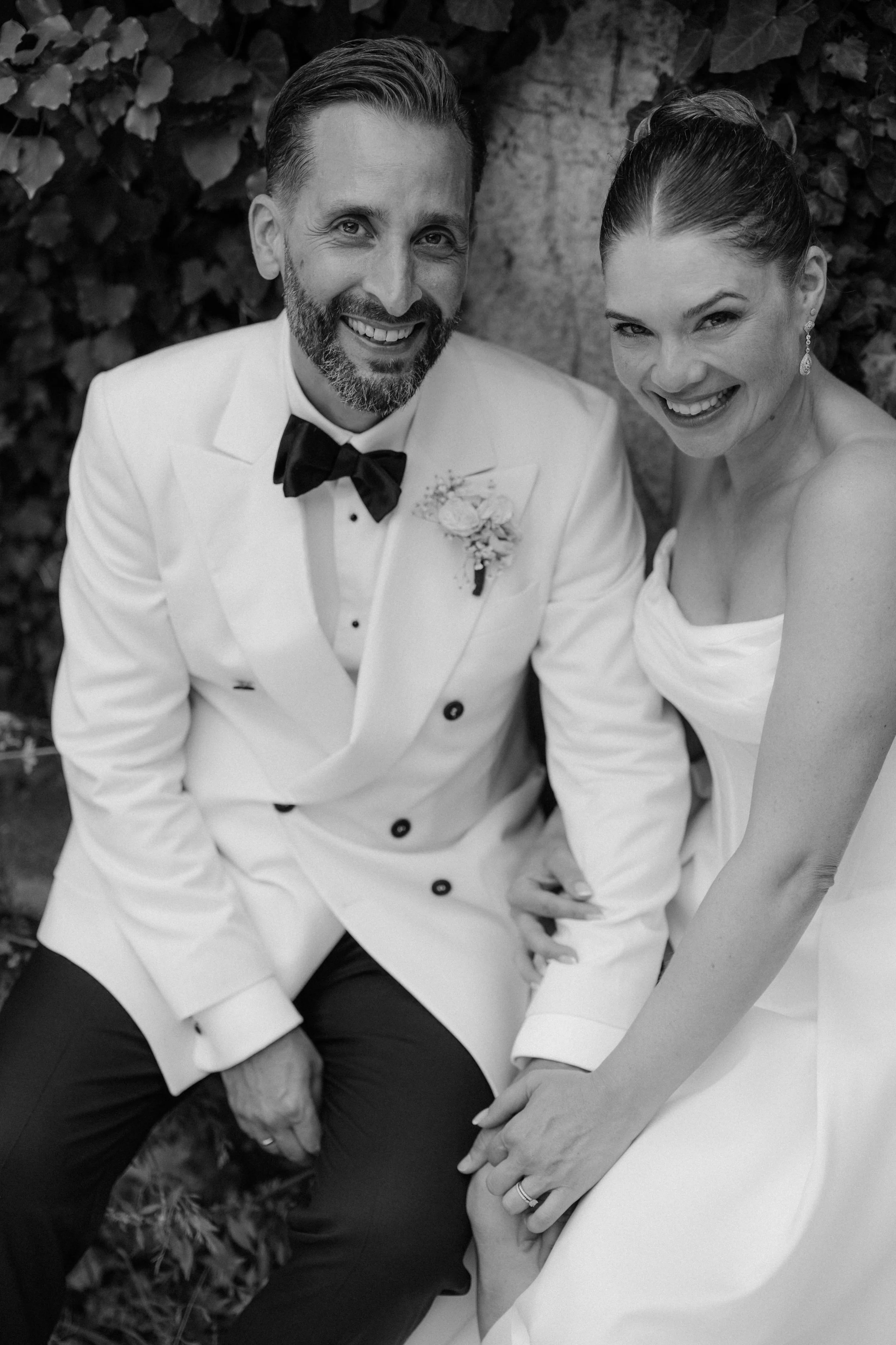 Black and white photo of a smiling man in a tuxedo and a woman in a wedding dress sitting outdoors with greenery behind them.