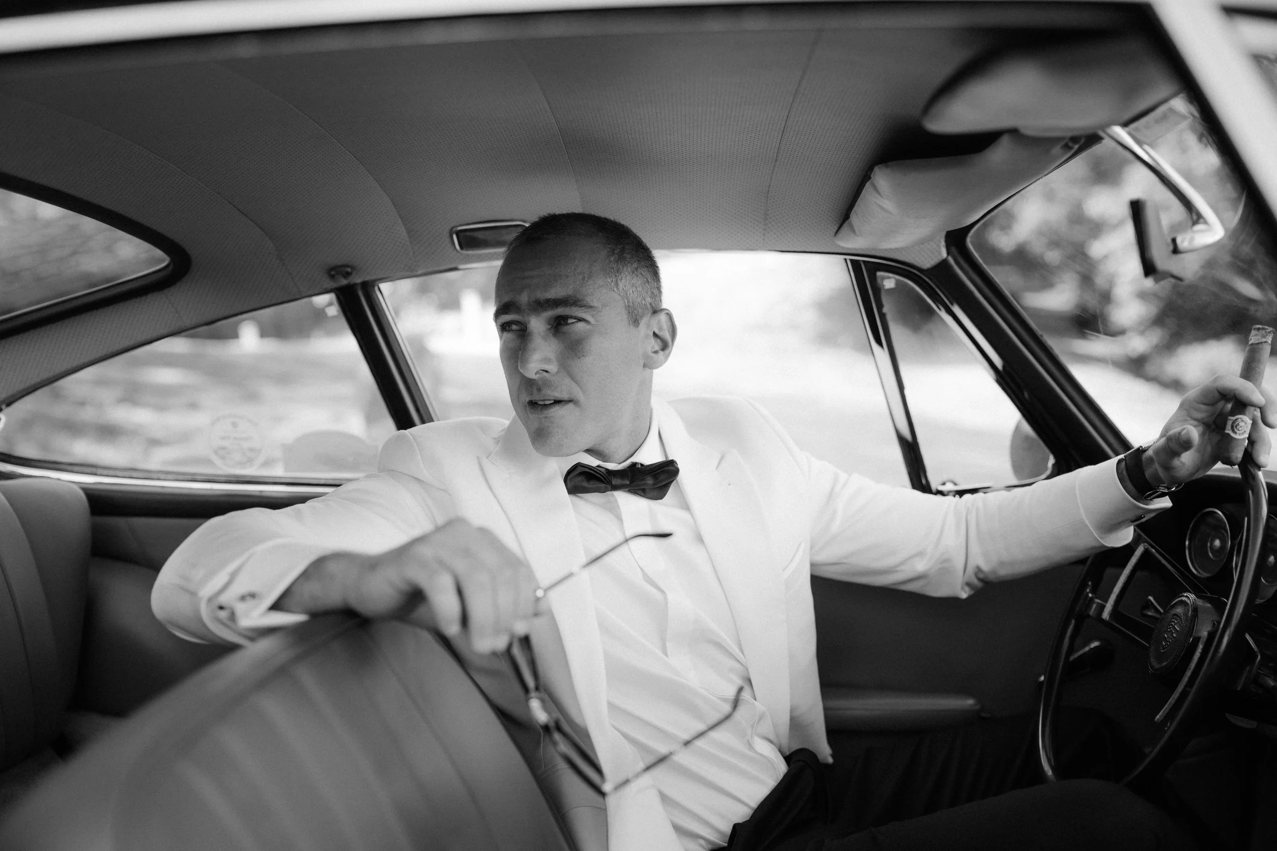A man in a tuxedo with a bowtie, sitting in the backseat of a car, holding a cigar in his right hand, looking to his left.