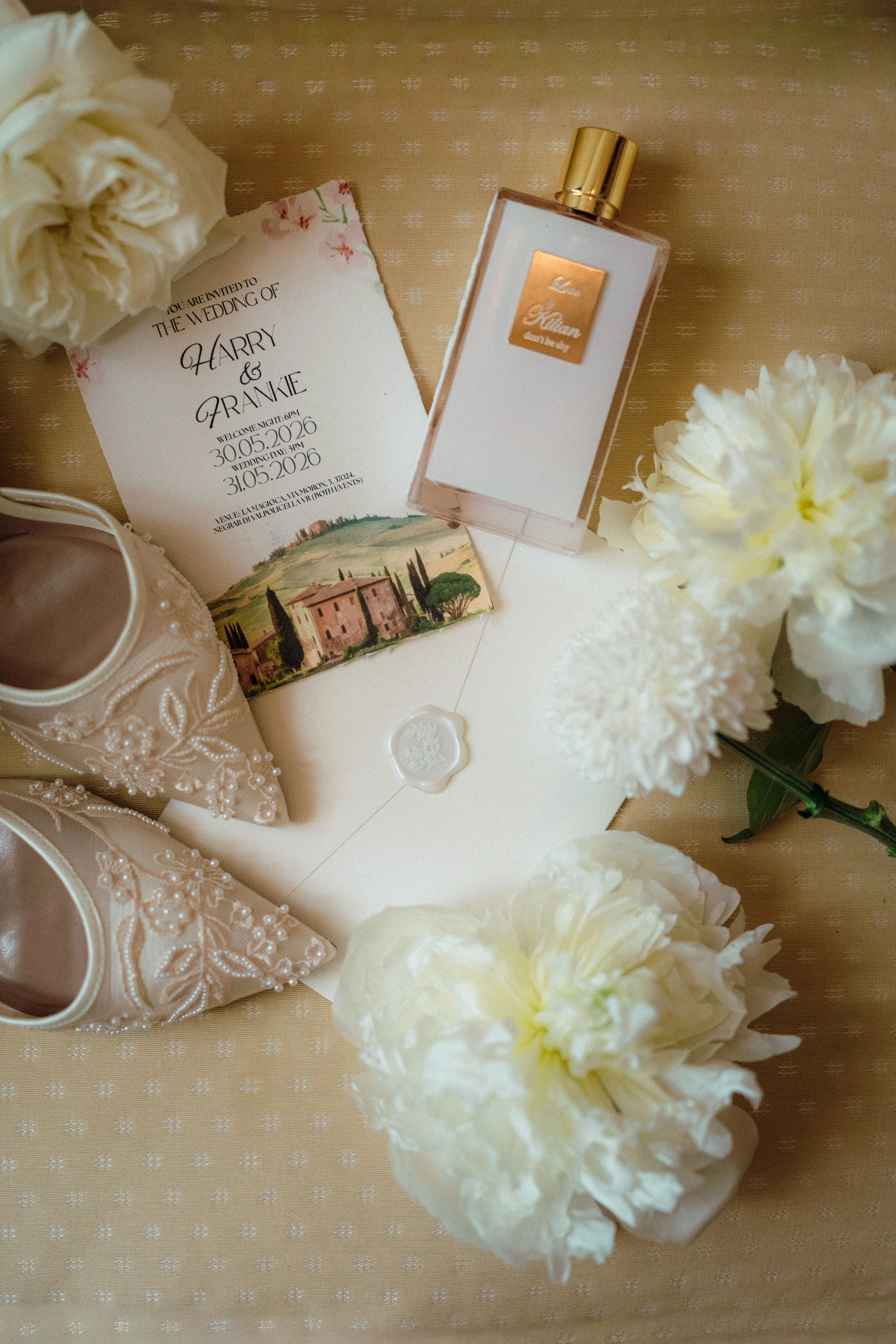 Wedding invitation card with floral design, a bottle of Love Kyliean perfume, a pair of beige shoes with pearl embellishments, and white flowers on a beige patterned tablecloth.
