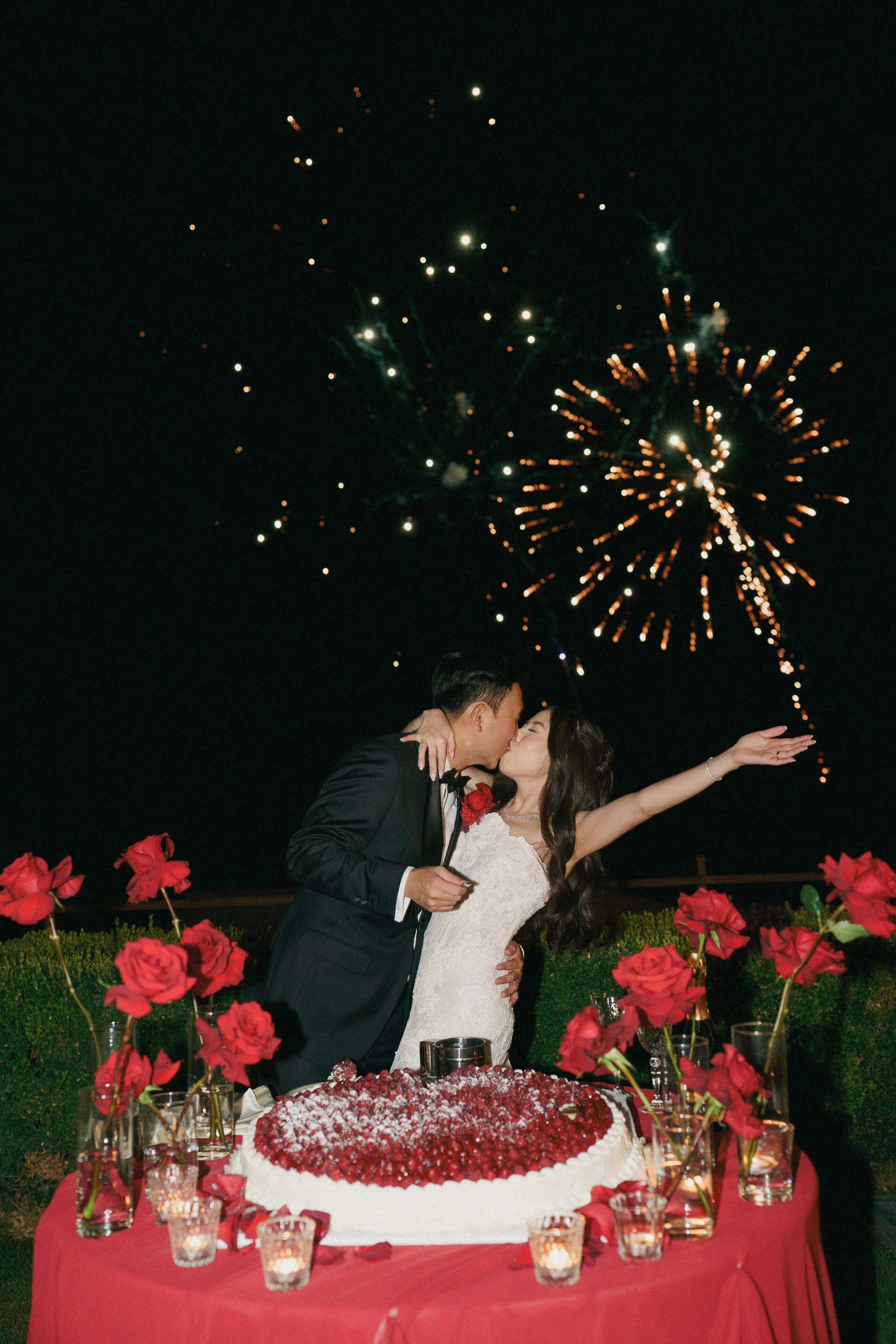 A newlywed couple kissing at their wedding reception outdoors at night, with fireworks lighting up the dark sky in the background. The table in front of them is decorated with red roses and candles.