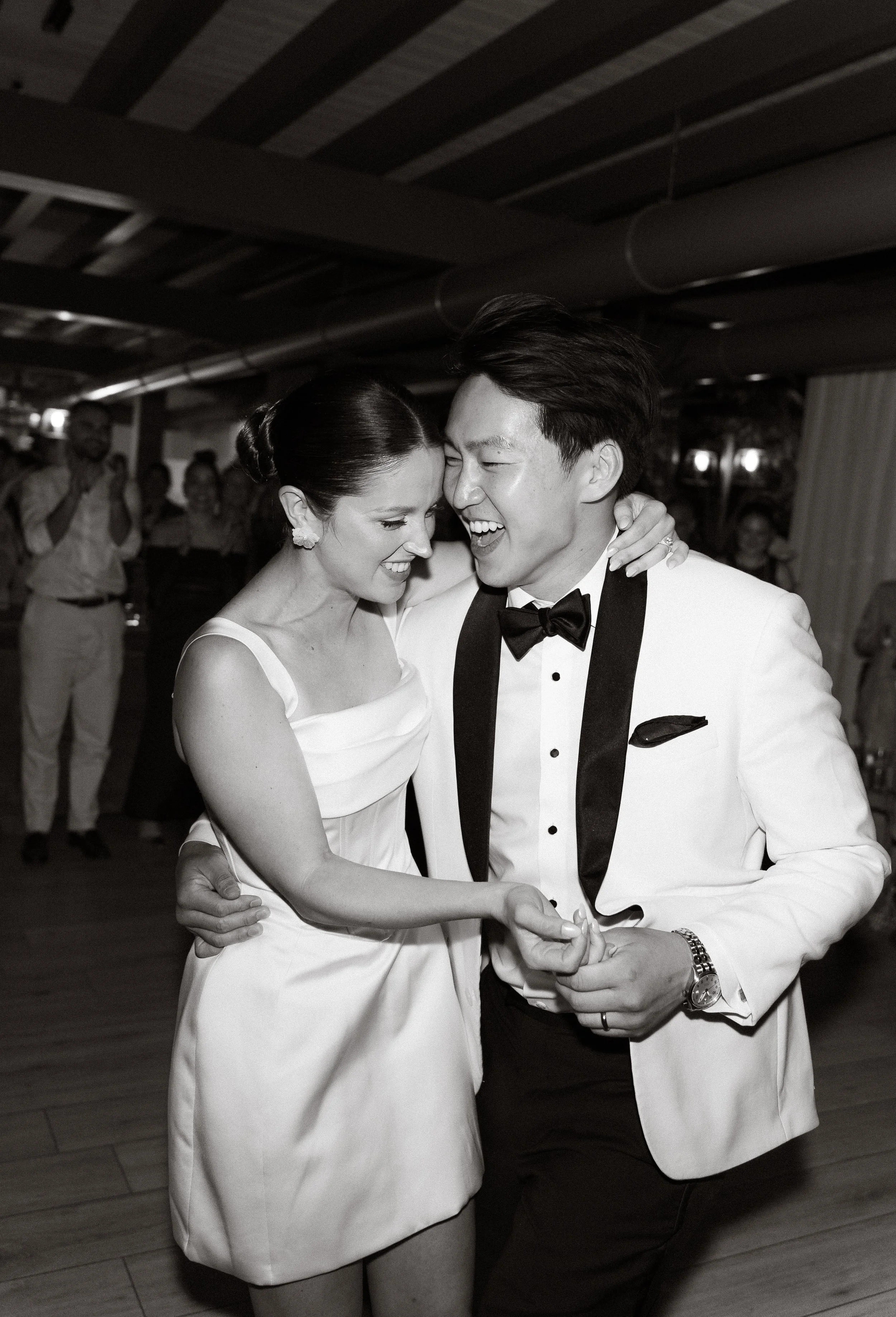 A couple dancing at a wedding reception, smiling and looking at each other, with guests in the background.