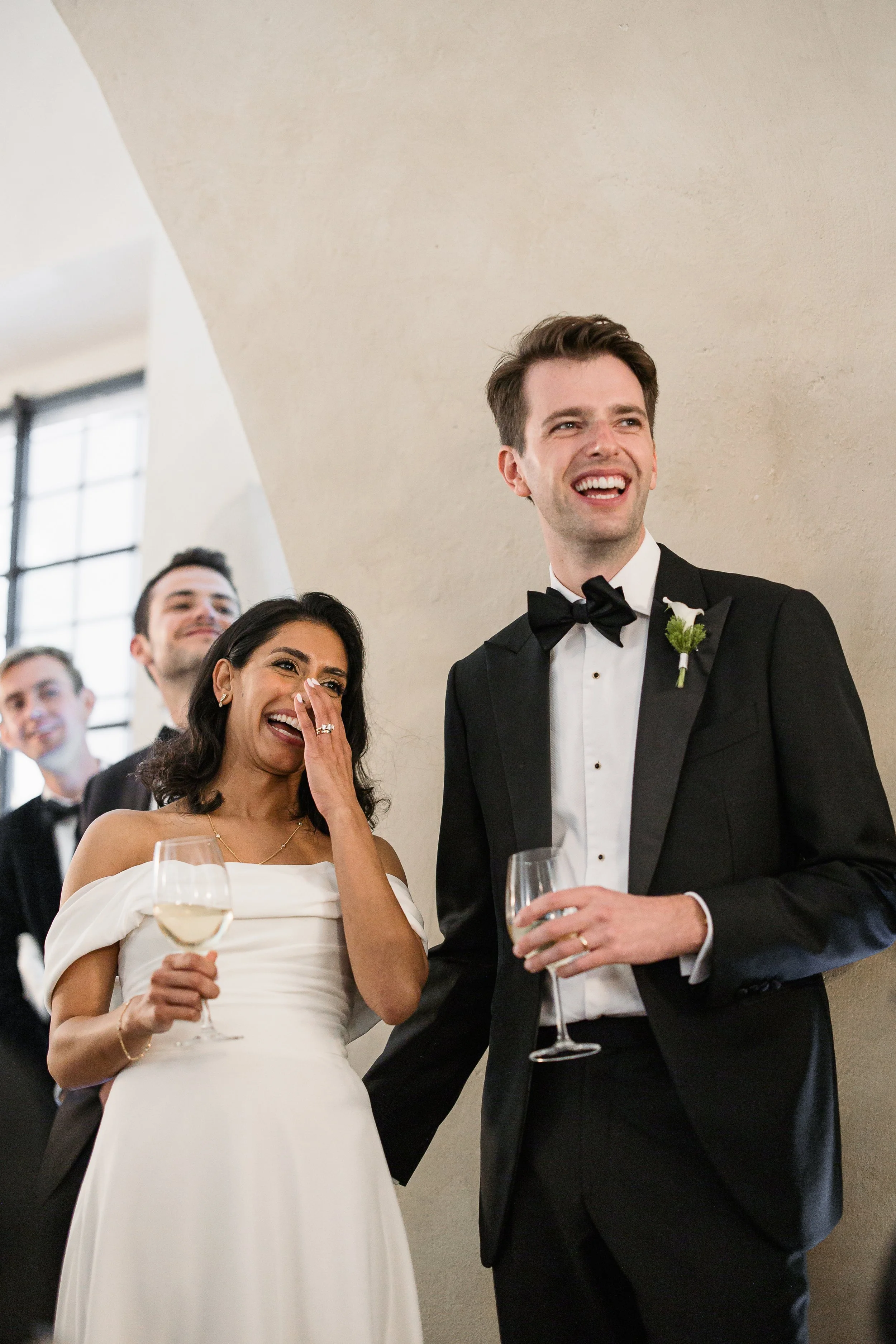 A smiling woman in a white dress and a woman in a tuxedo holding wine glasses at a wedding reception.