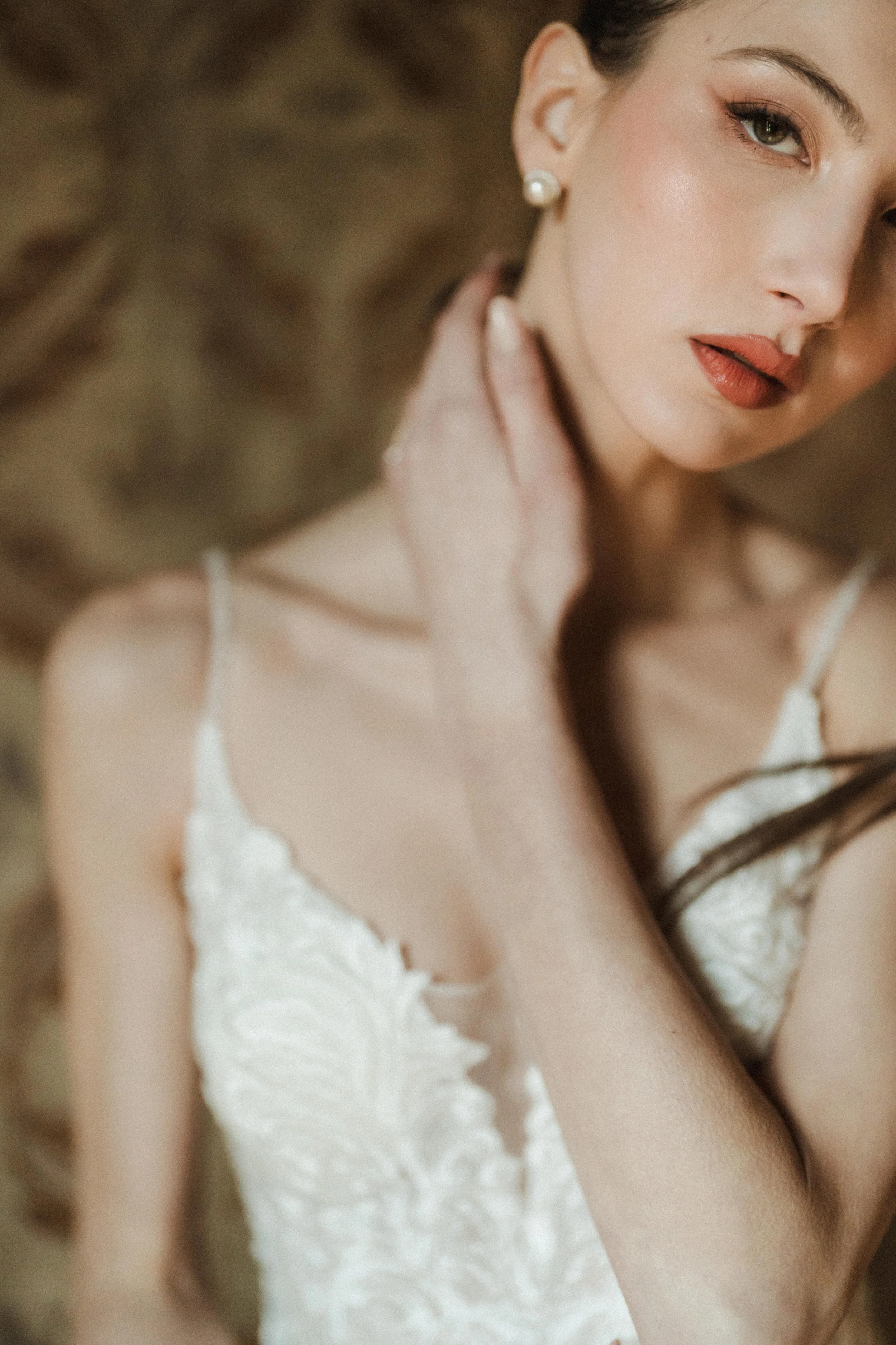 A close-up of a woman with light skin and makeup, wearing pearl earrings and a white lace top, touching her neck with her hand, in a soft focus photograph.