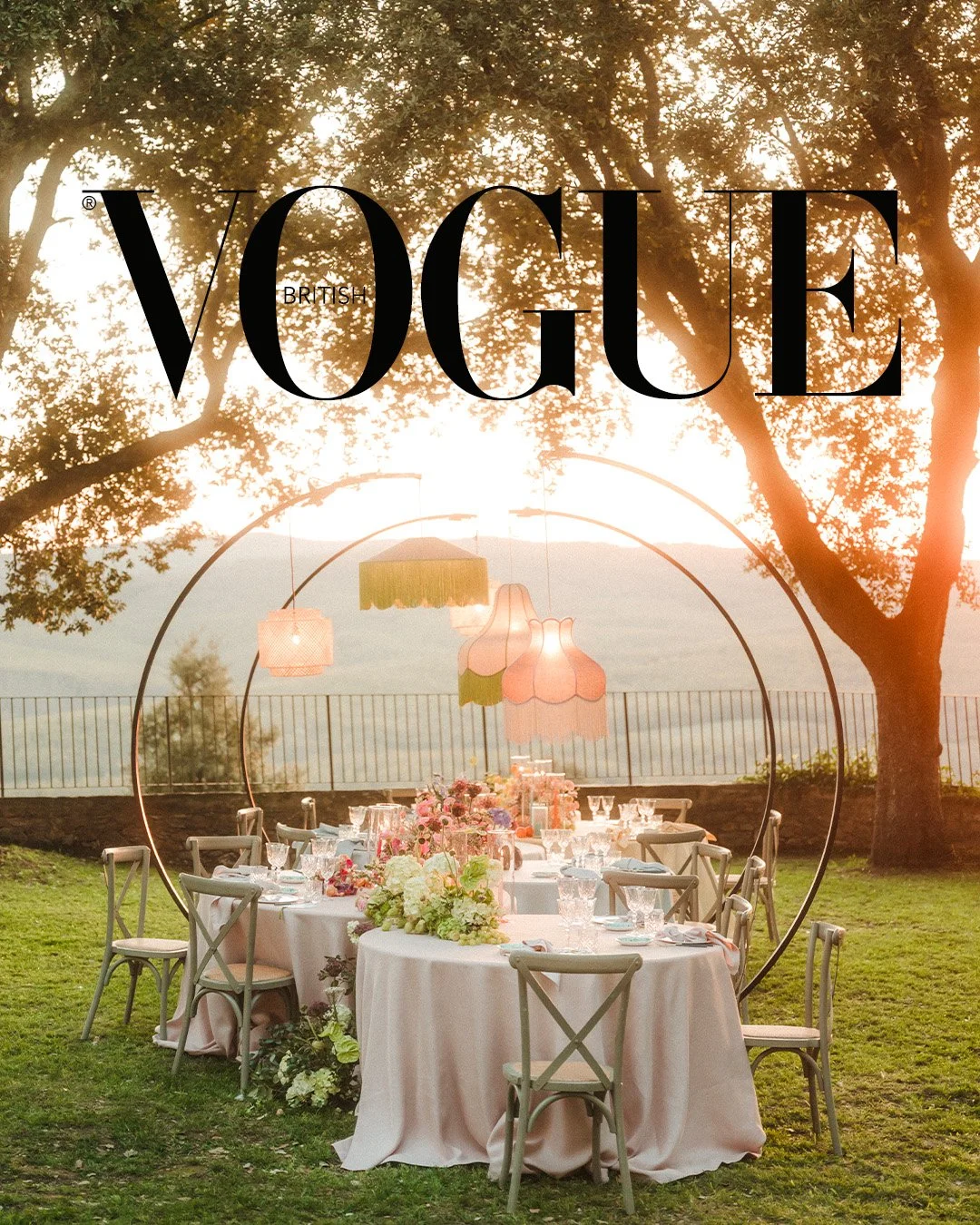 A beautifully decorated outdoor dining setup with a long table covered with a white tablecloth, elegant glassware, and floral centerpieces, set beneath hanging pendant lights. Large trees with a fence and scenic view in the background, in a sunset or sunrise lighting. Overlaid with the 'Vogue British' logo.