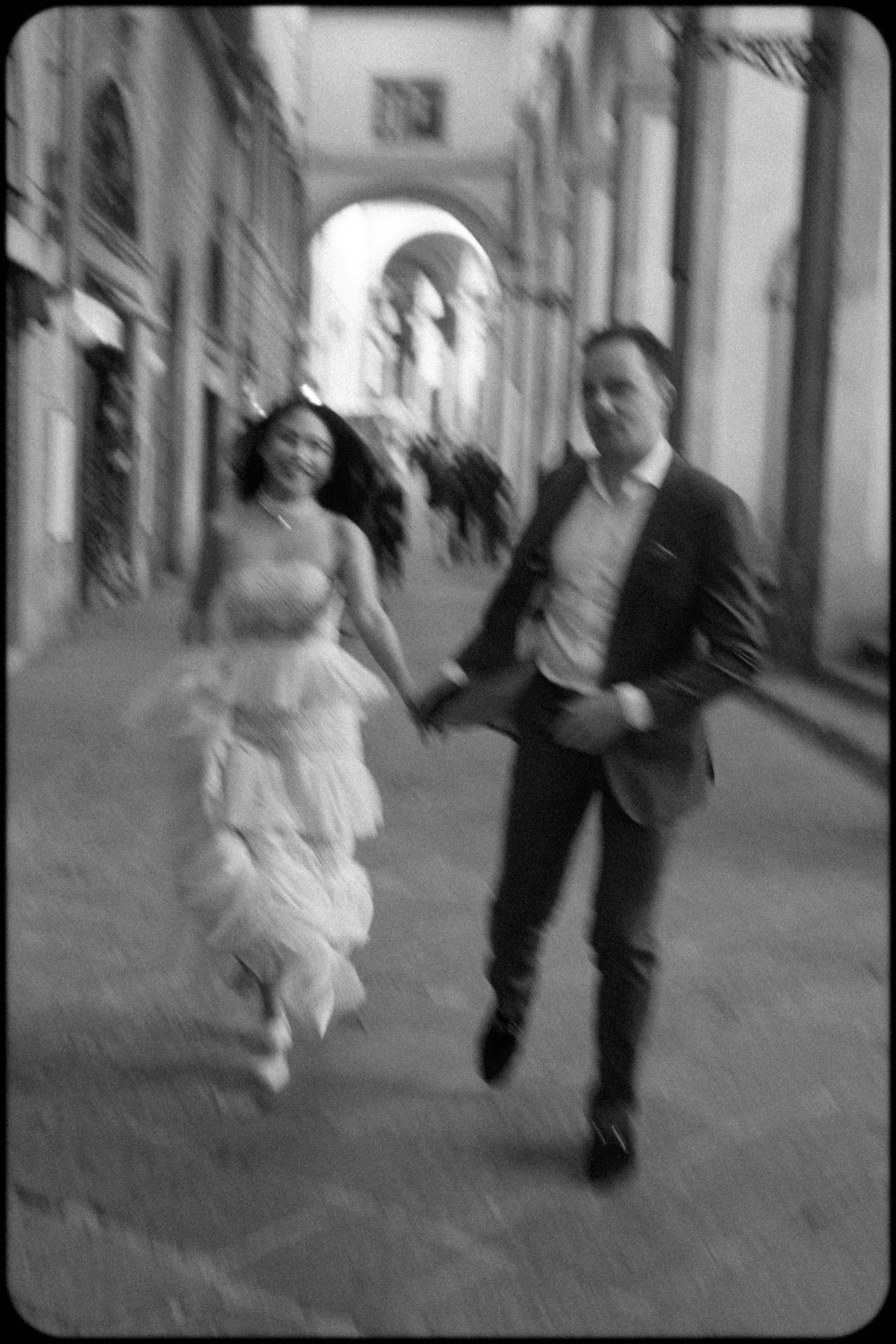 A woman in a fancy strapless dress and a man in a dark suit and dress shirt are dancing in a decorated hallway with statues and ornate architectural features.