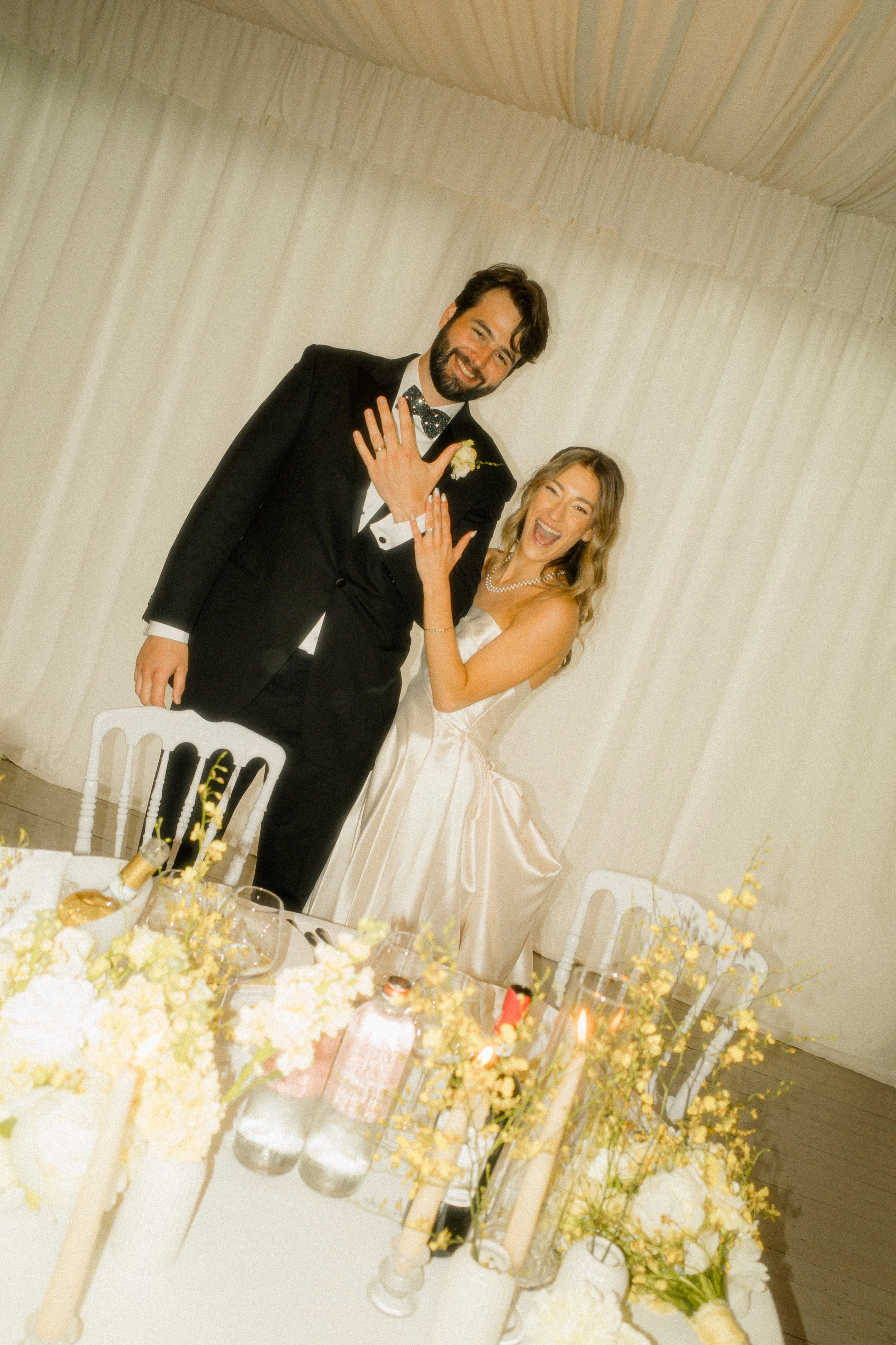 A man and woman dressed in formal wedding attire smiling and showing their wedding rings at a wedding reception table decorated with yellow and white flowers and various drinks.