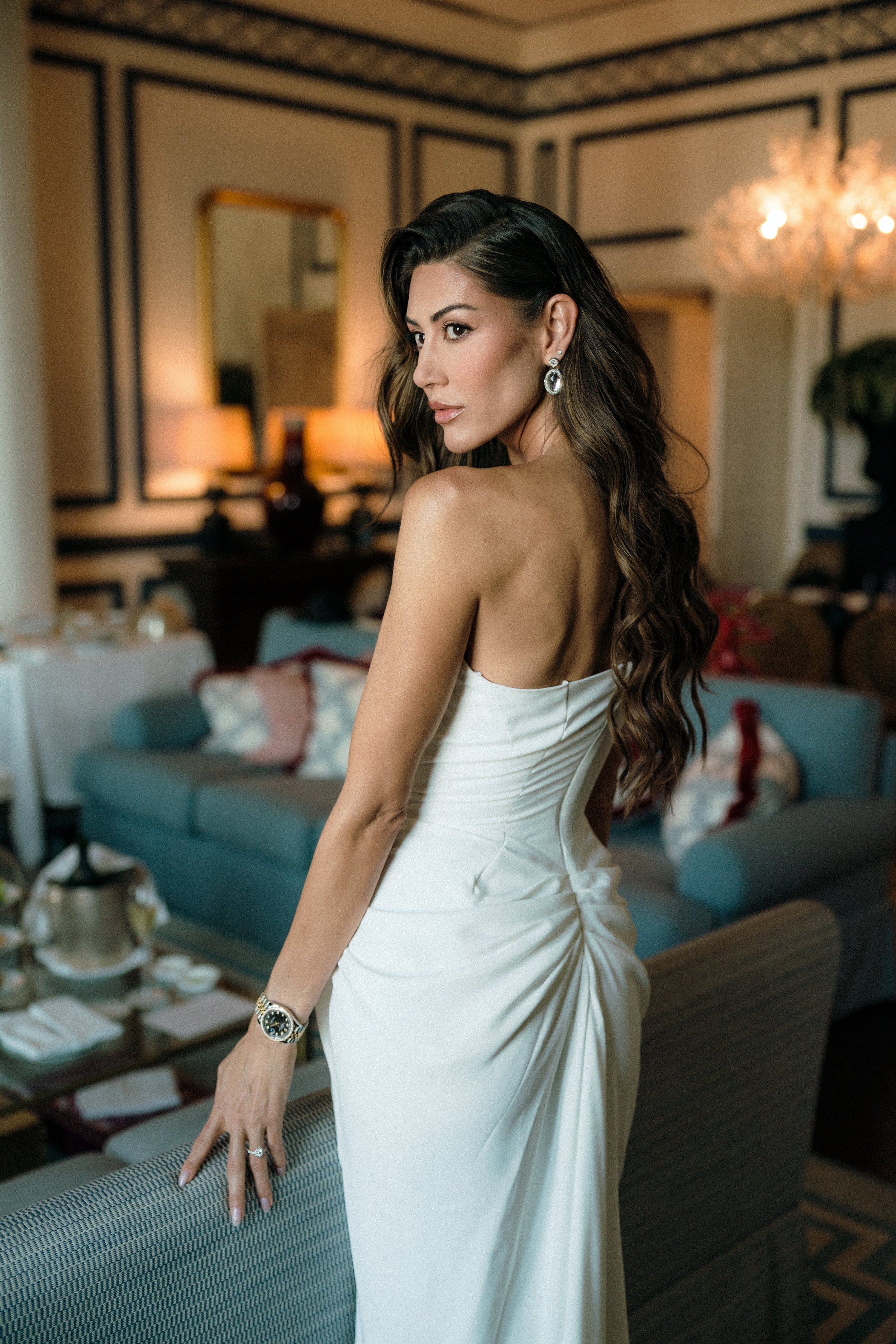 A woman with long, wavy brown hair in a white strapless dress, wearing earrings and a watch, standing in a living room with decorated walls, lamps, and furniture.