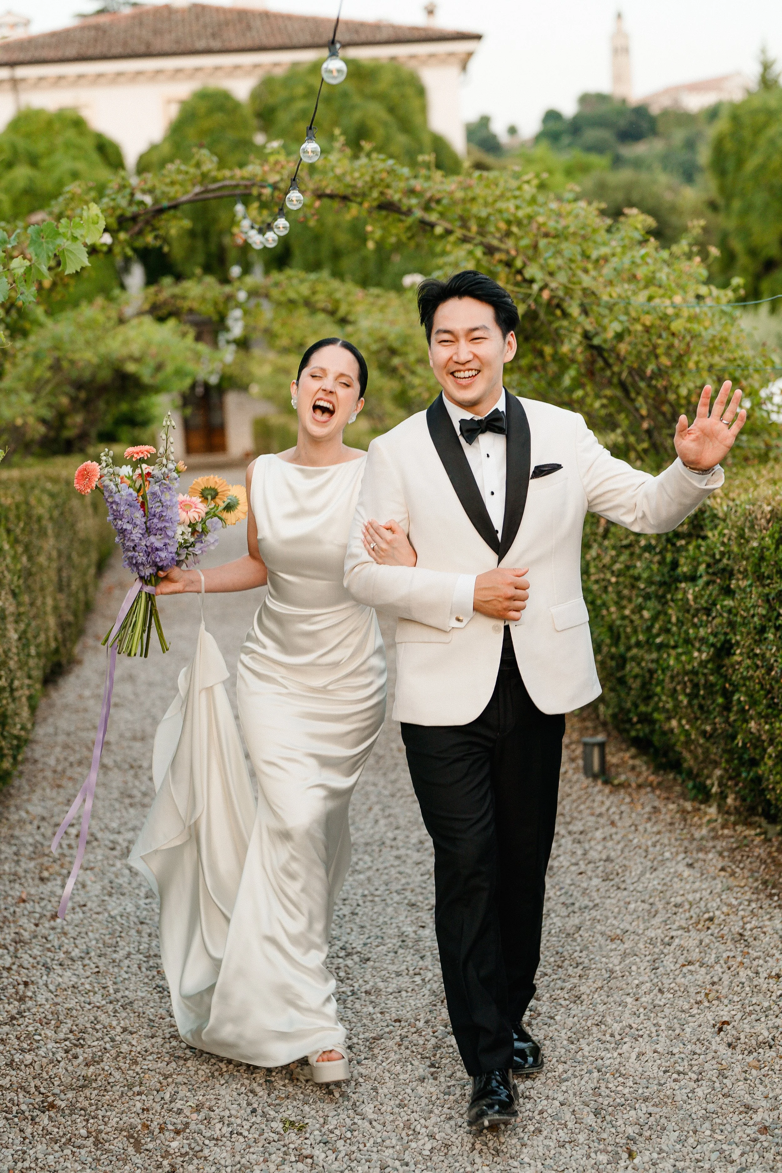A bride and groom walking arm-in-arm outdoors, smiling and laughing. The bride is holding a bouquet of flowers, wearing a white wedding dress, and the groom is dressed in a white tuxedo jacket with black pants and a bow tie. The background features g