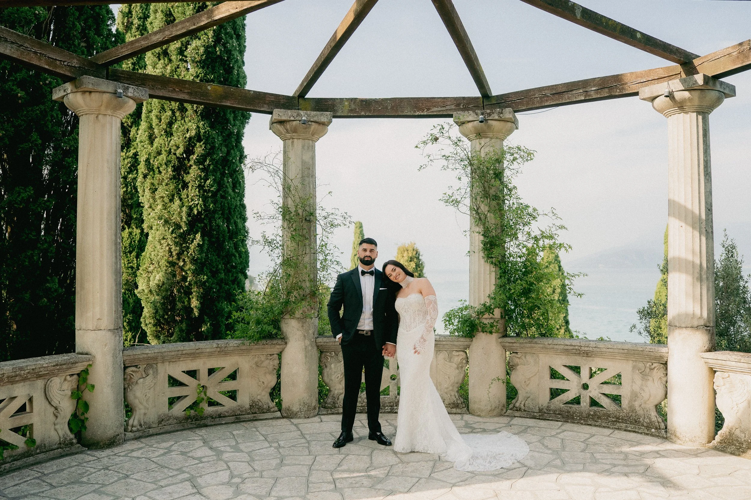 A bride and groom standing together outdoors under a stone balcony with columns, surrounded by trees and greenery.