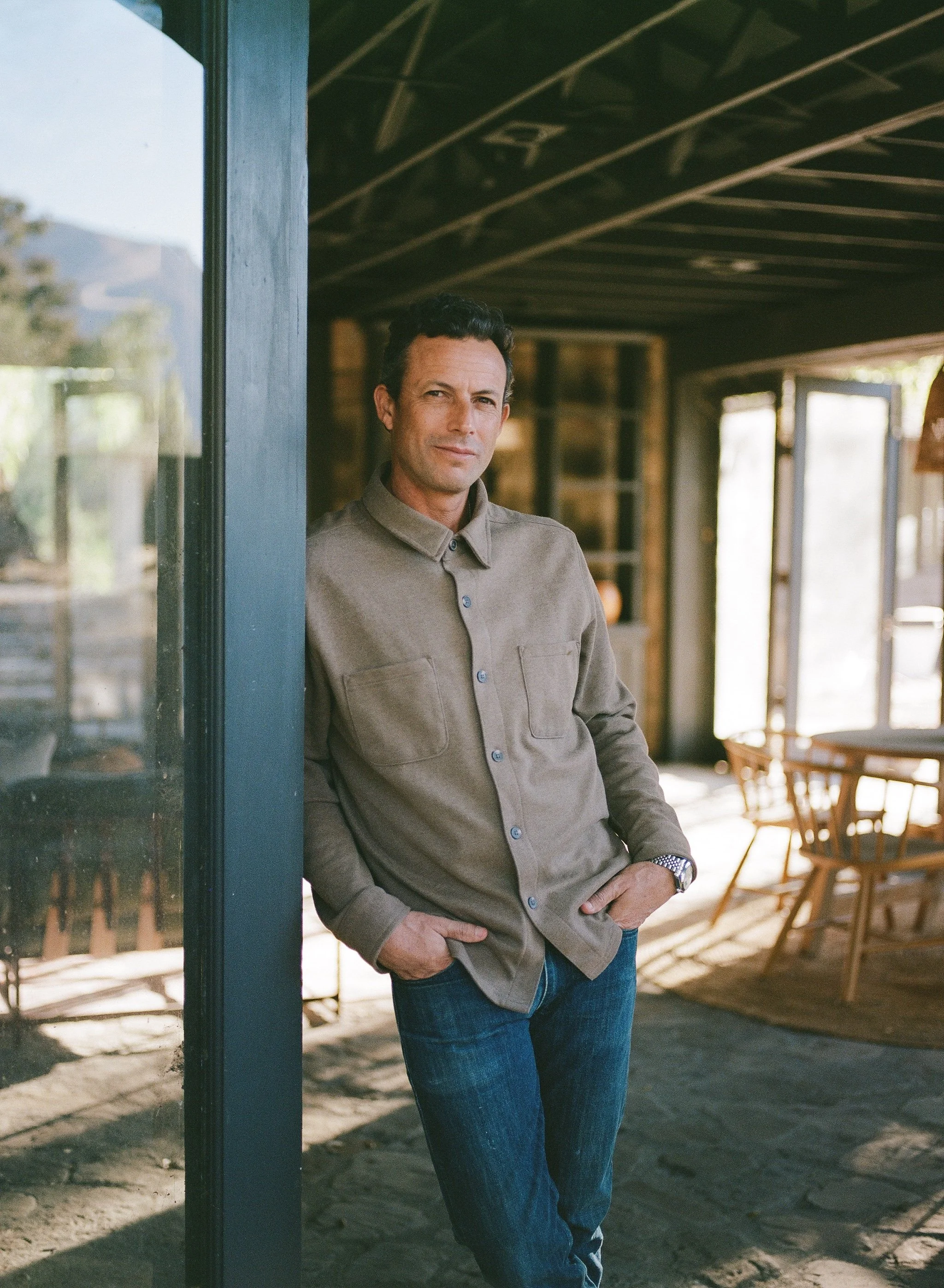 A man standing indoors near a window and door, wearing a khaki long-sleeved shirt and jeans, with his hands in his pockets, in a rustic setting with natural light.